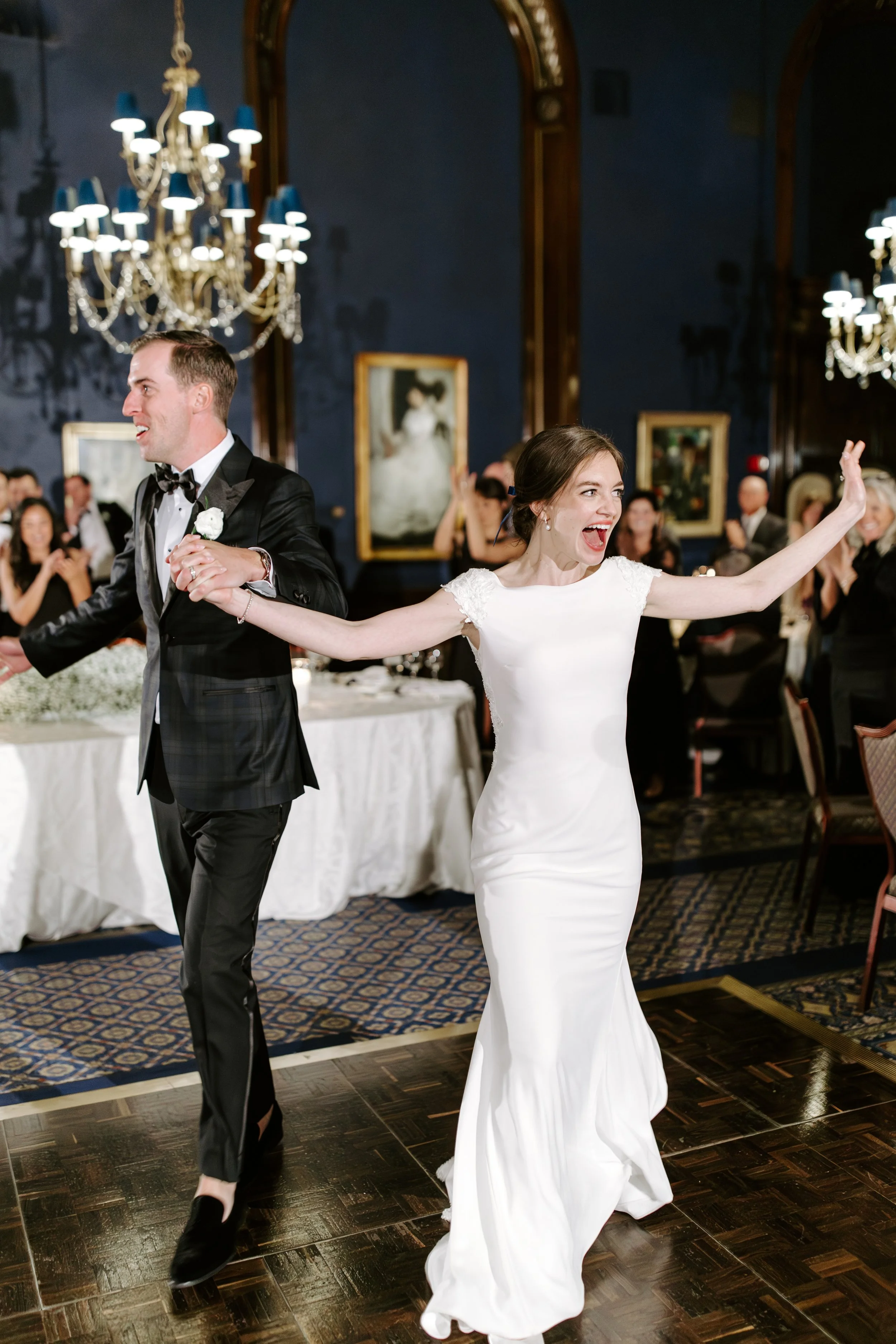 A bride and groom dancing at a wedding reception in a decorated hall with chandeliers, paintings, and guests clapping in the background.