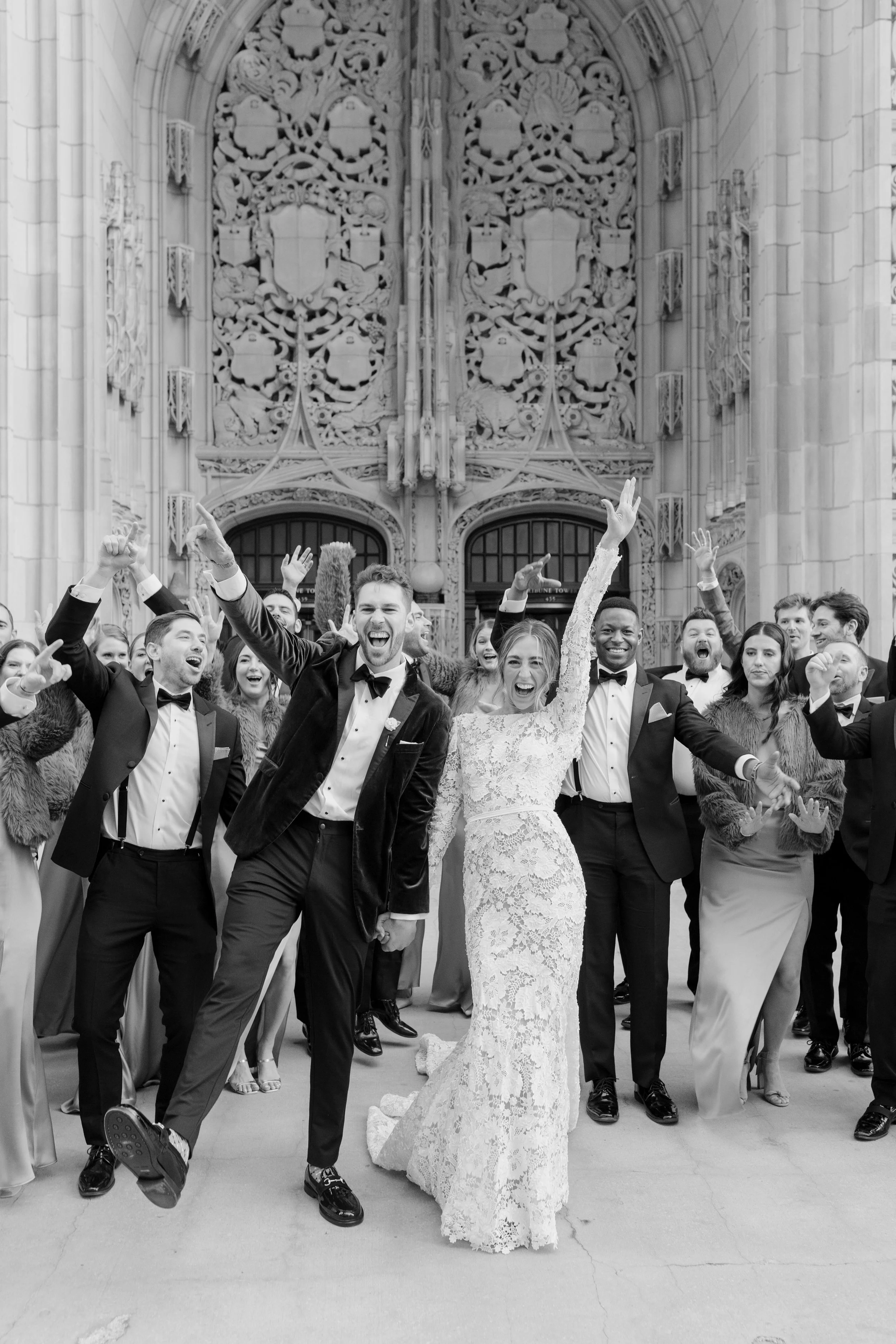 A group of wedding guests in formal attire, smiling and celebrating in front of a Gothic-style church entrance with ornate carvings.