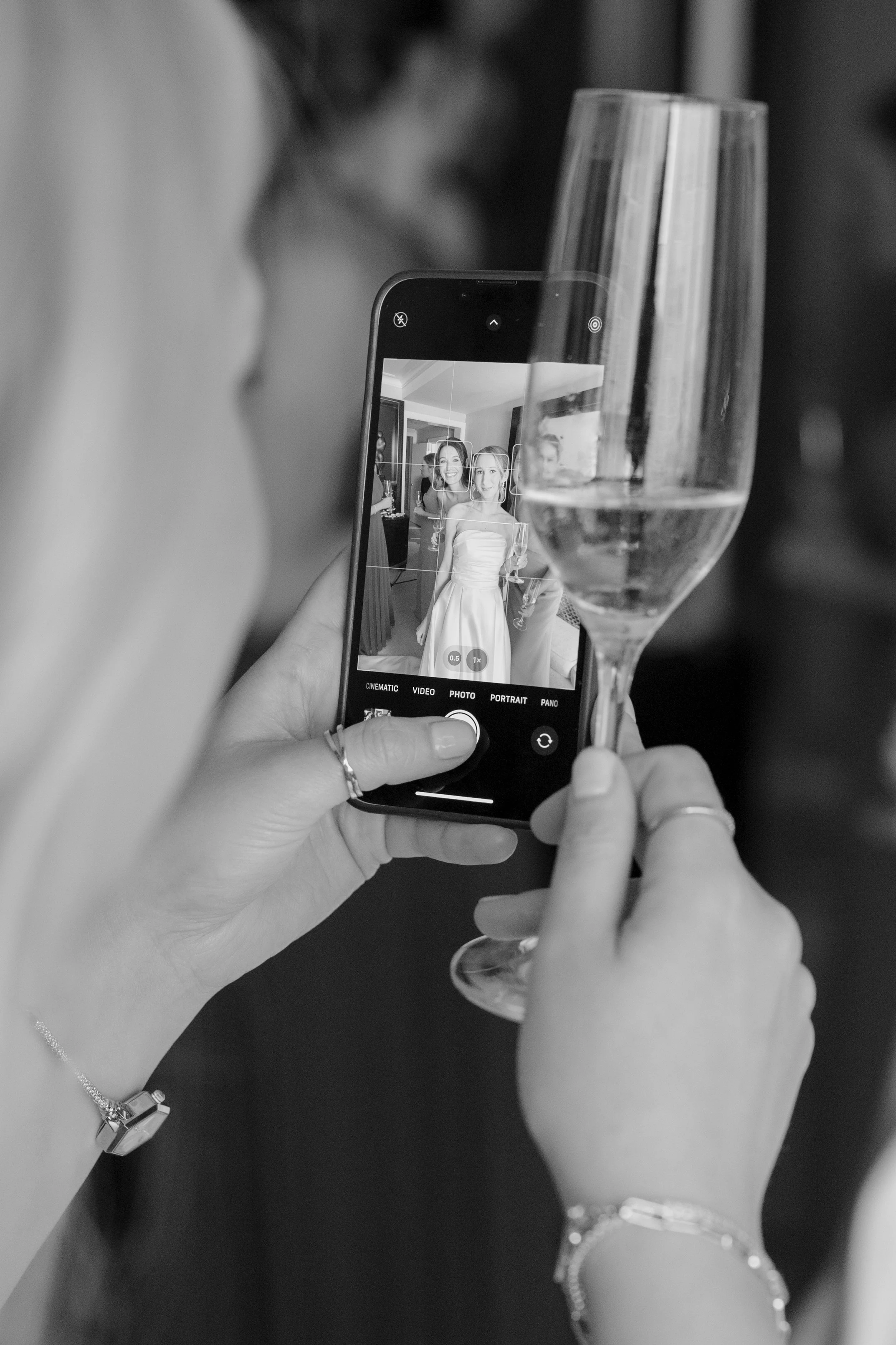 Person taking a photo of a bride through a smartphone at a celebration, with a glass of wine in the foreground.