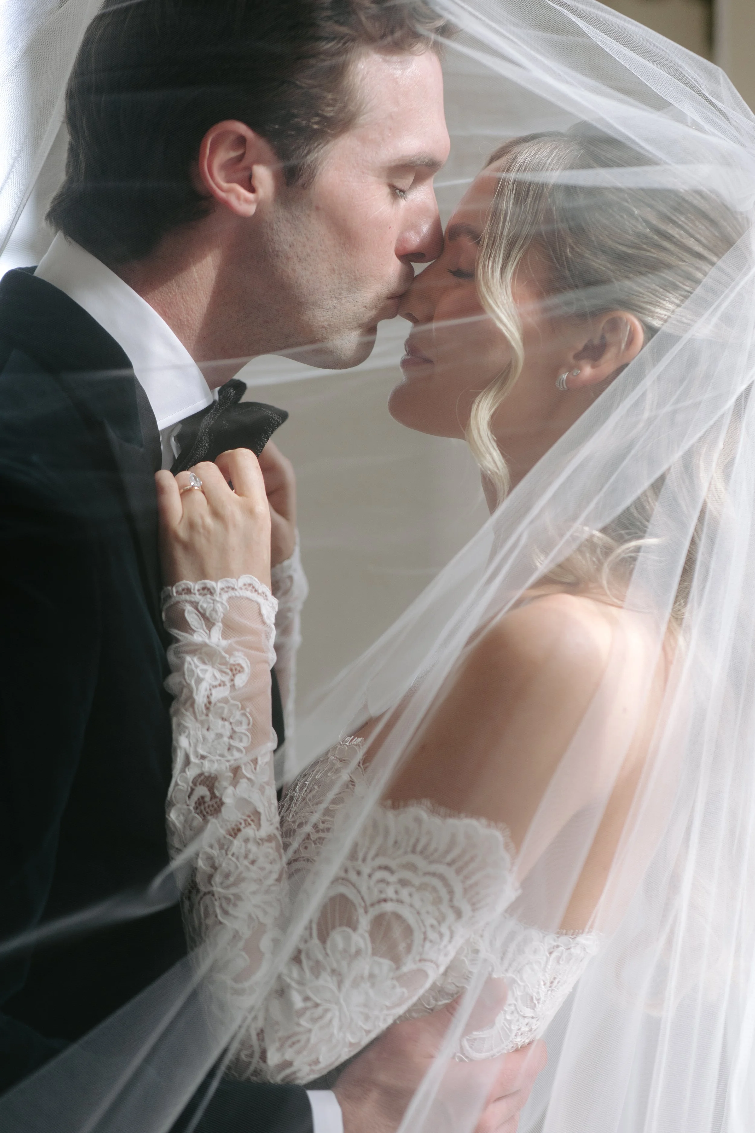 A bride and groom sharing a kiss under a wedding veil, with the bride wearing lace and the groom in a tuxedo.