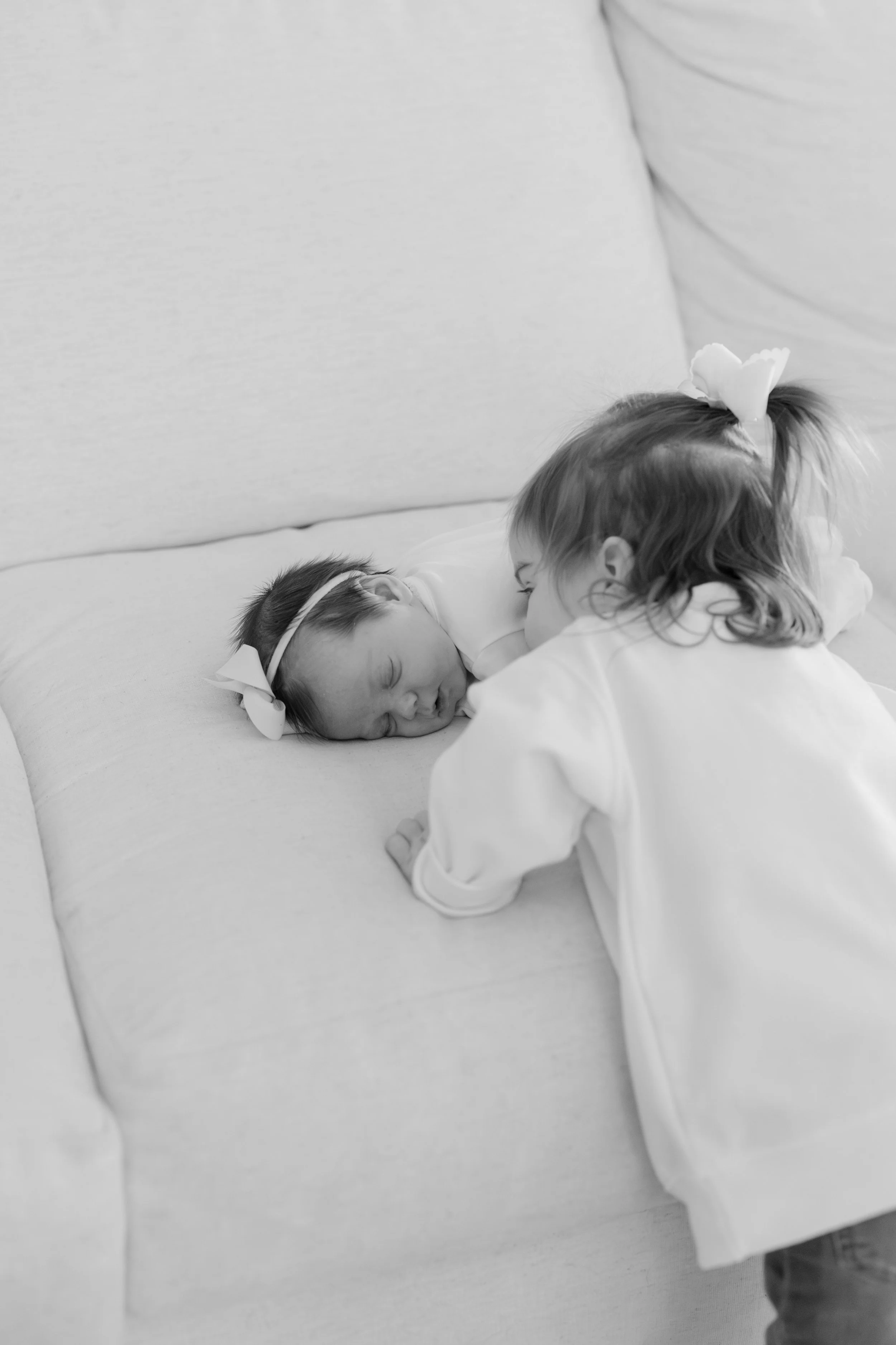 A woman is leaning over a newborn baby who is lying on a pillow on a bed. The baby is wearing a headband with a bow. The woman appears to be gently kissing or inspecting the baby's face.