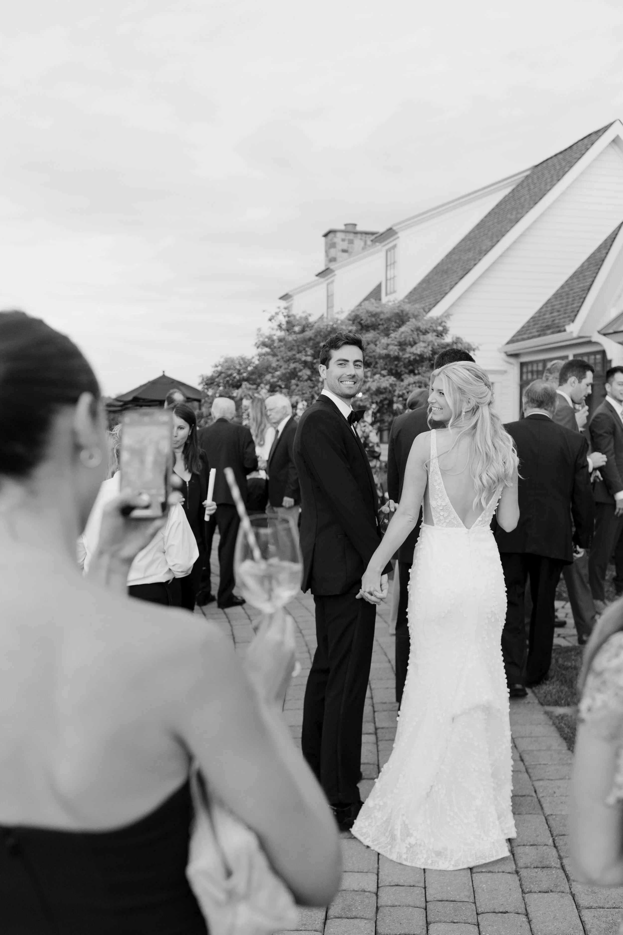 A bride and groom holding hands at an outdoor wedding reception with guests and a house in the background.