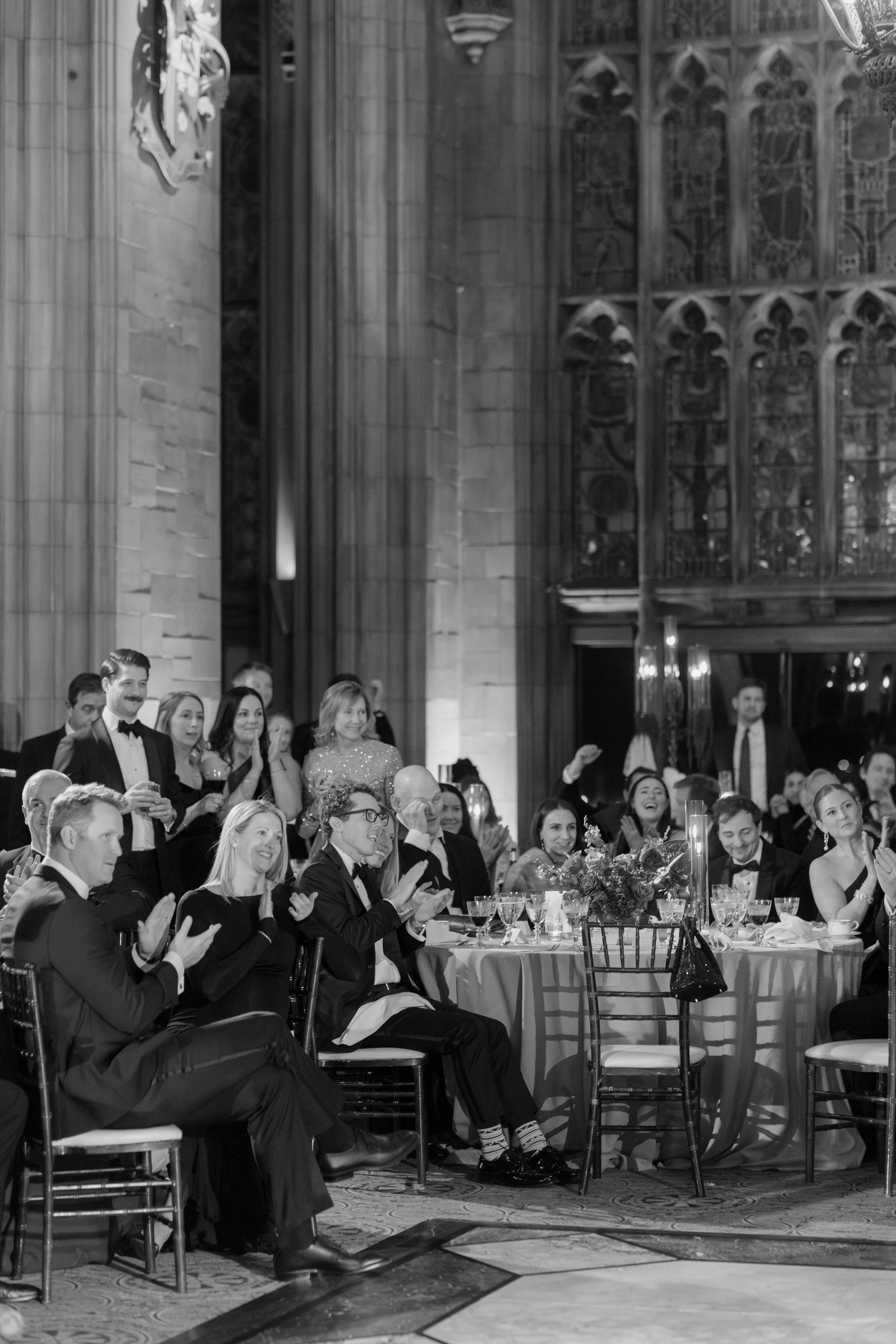 People dressed in formal attire attending a banquet or reception in a grand hall with Gothic architectural features and stained glass windows, seated at decorated tables.