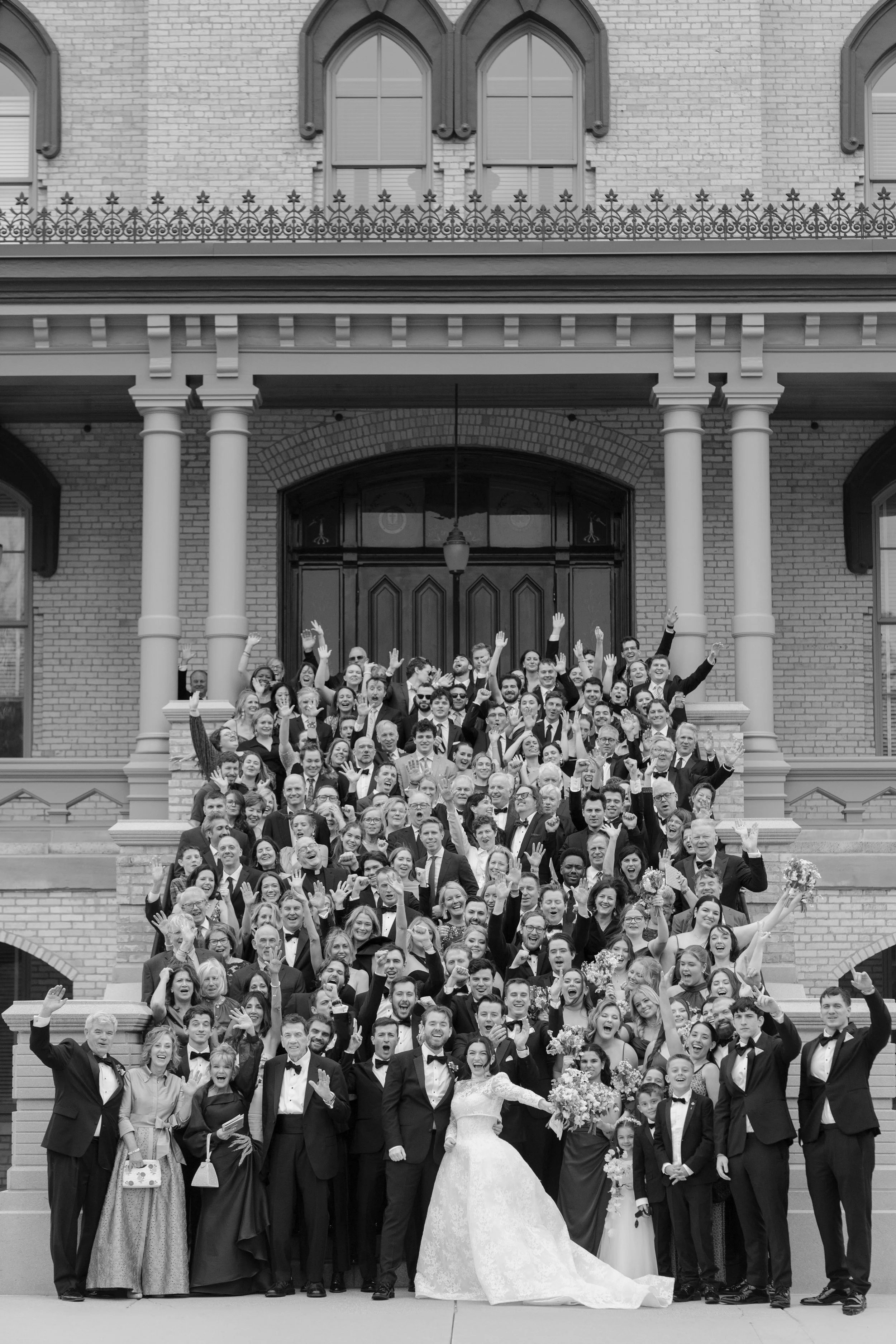 A large group of people dressed in formal attire, including tuxedos and gowns, gathered on the steps of a brick building with large windows, celebrating a wedding. The bride in a white gown and veil is at the front, surrounded by friends and family w