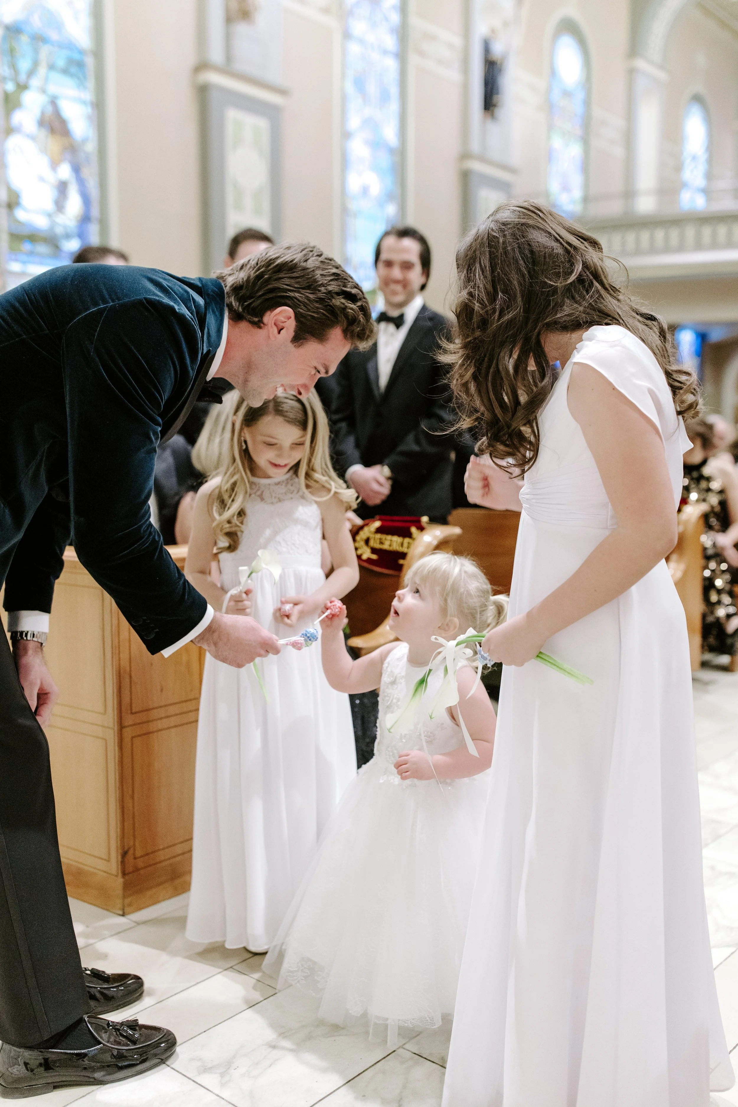 A wedding ceremony inside a church with two young flower girls in white dresses, a woman in a white dress, and men in suits. The woman and flower girls are holding flowers, and one man appears to be giving a blessing or saying a prayer. The backgroun