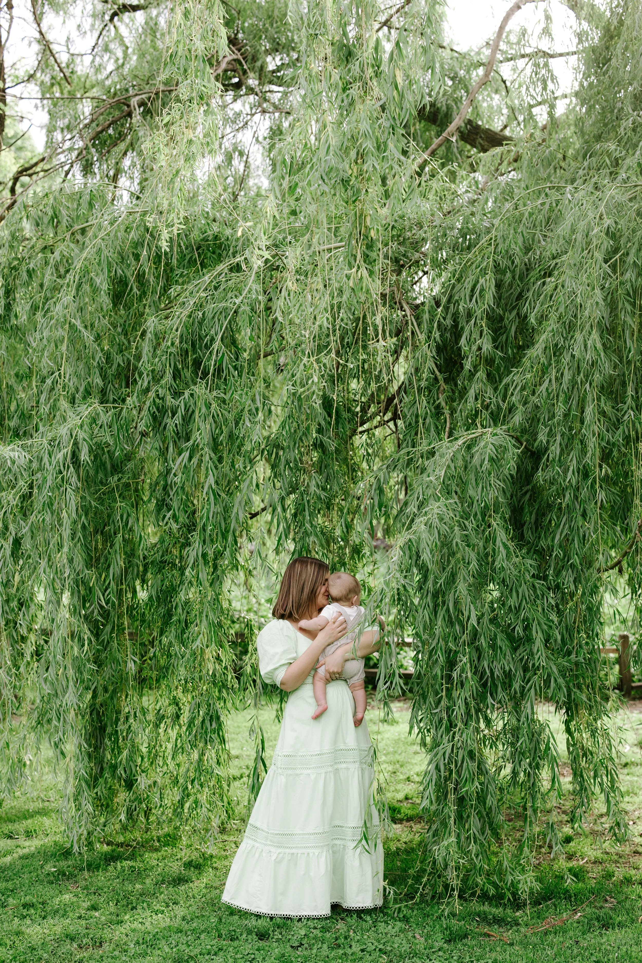 A woman in a white dress holding a baby near a tall weeping willow tree with lush green leaves.