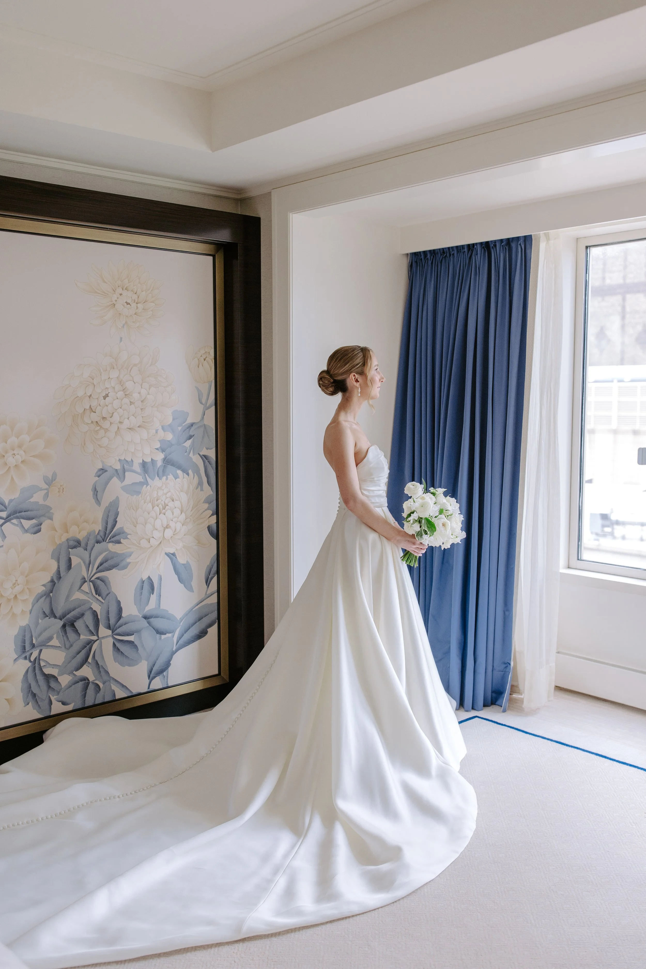 A bride in a white wedding dress holding a bouquet of white flowers, standing in a room with blue curtains and floral artwork on the wall.