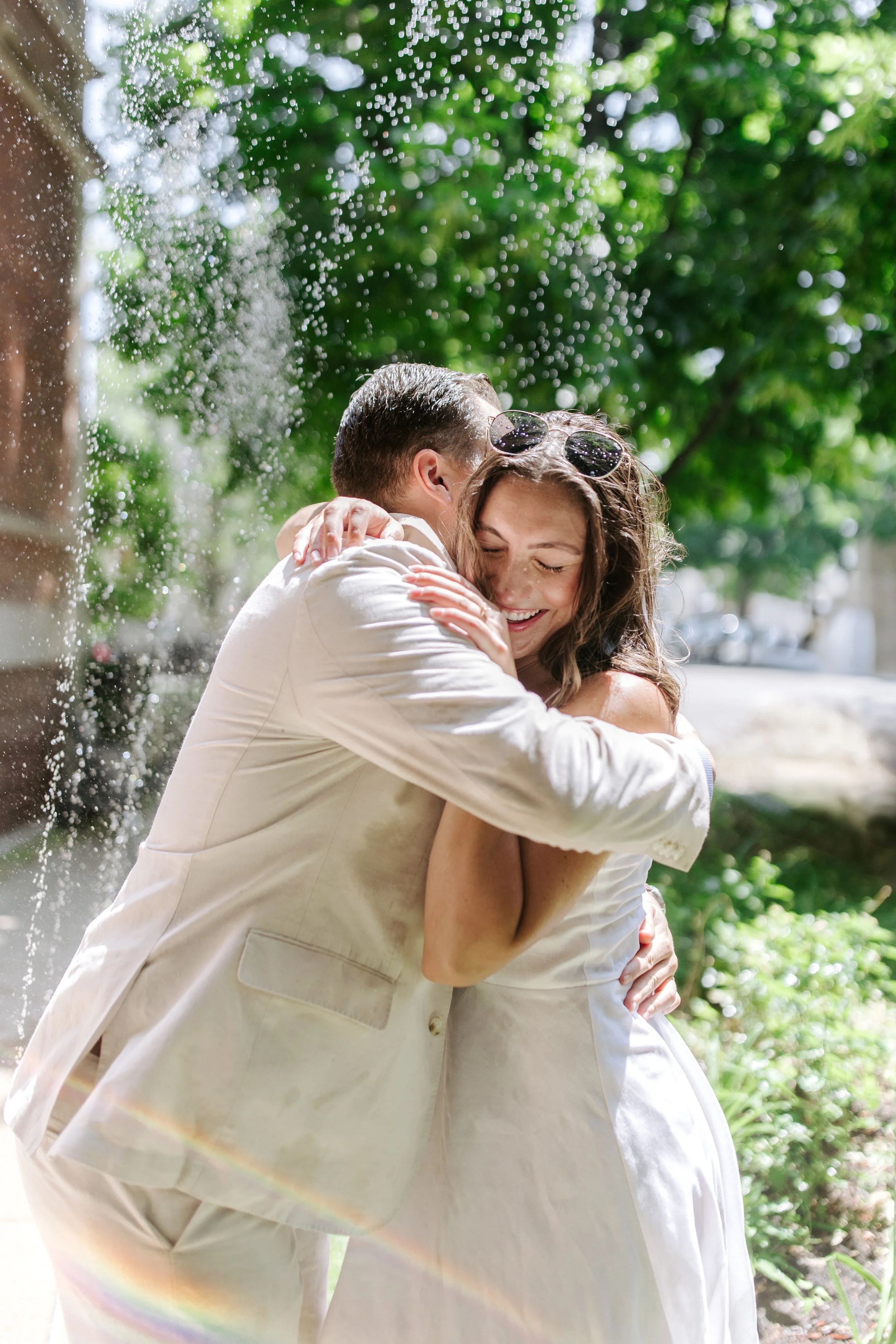 A couple is hugging outdoors on a sunny day, with the woman smiling and the man with his face close to her. They are dressed in white and have sunglasses on their heads. The background features green trees and dappled sunlight.