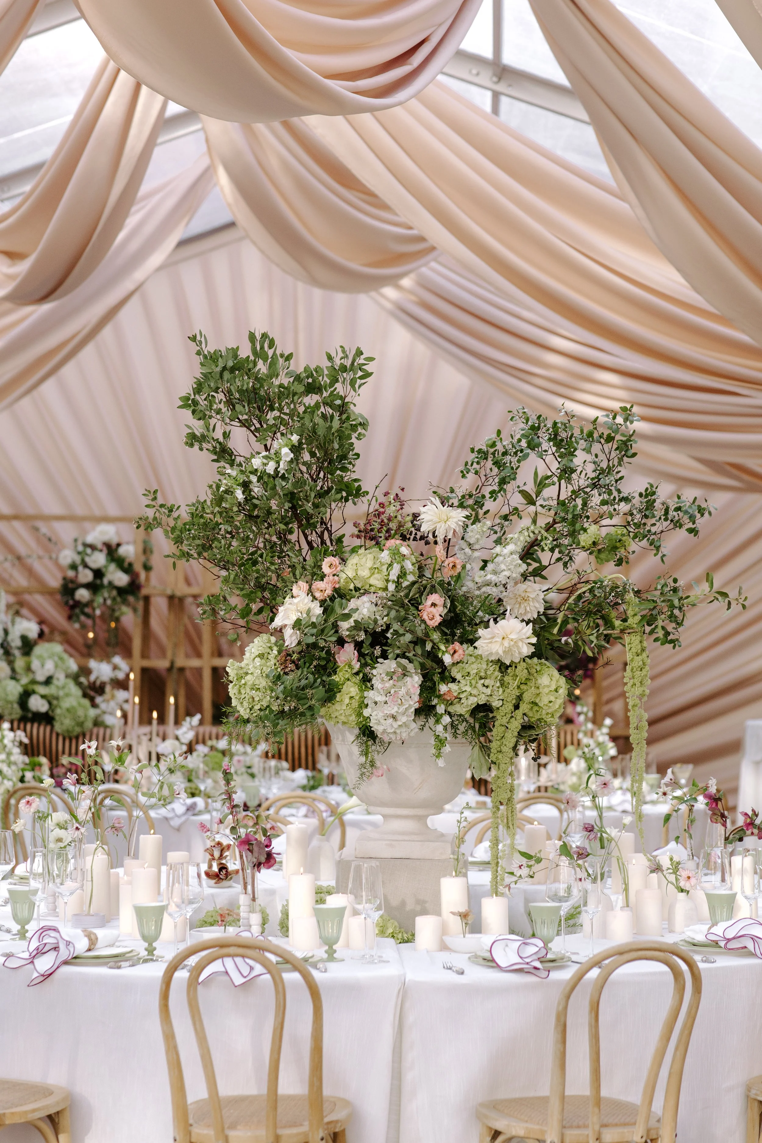 Elegant banquet table with a large floral centerpiece in a white urn, surrounded by white candles and tableware, under a cream-colored draped fabric ceiling.