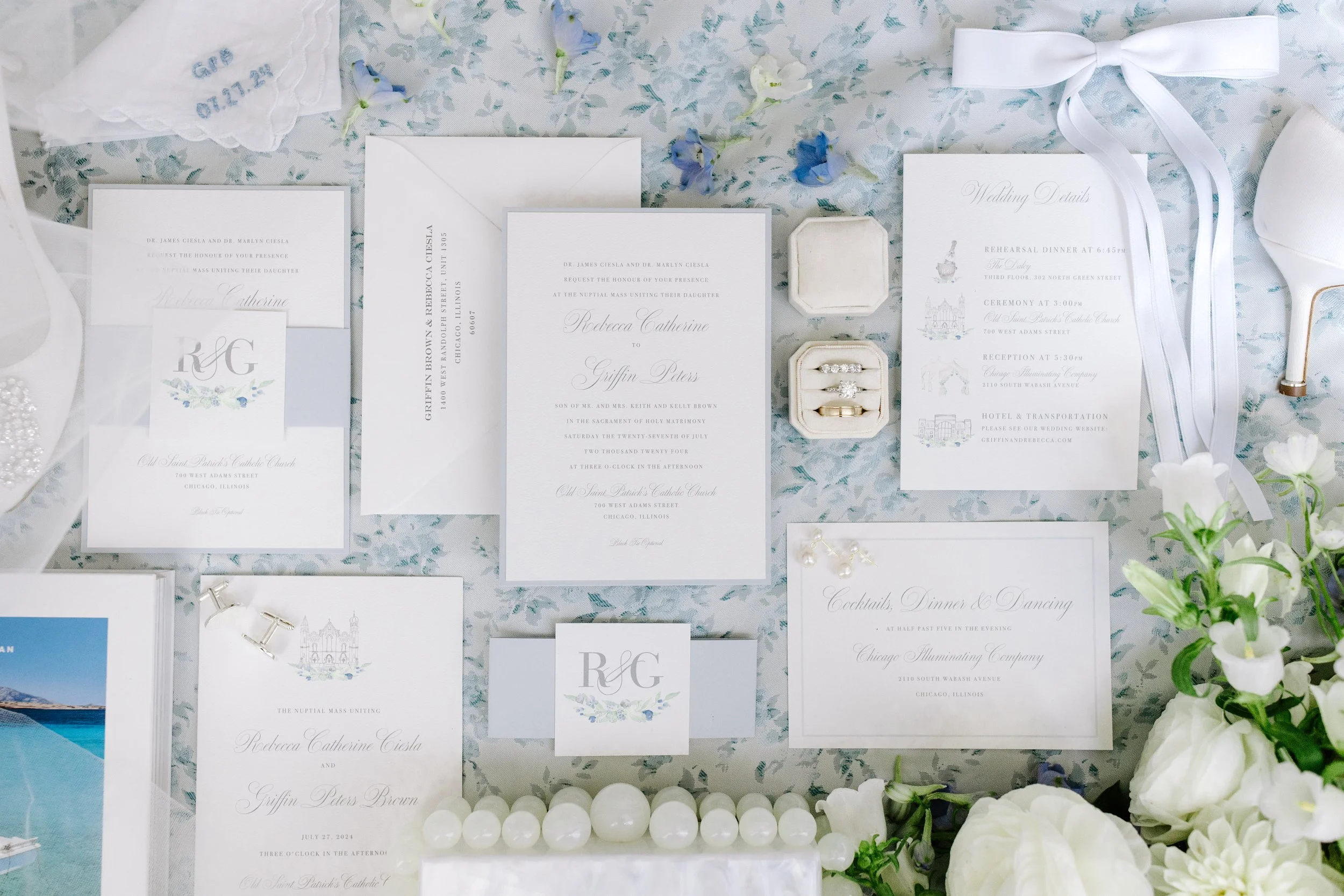 Wedding invitation suite with floral details, wedding rings in a white box, and scattered blue and white flowers on a light-colored surface.