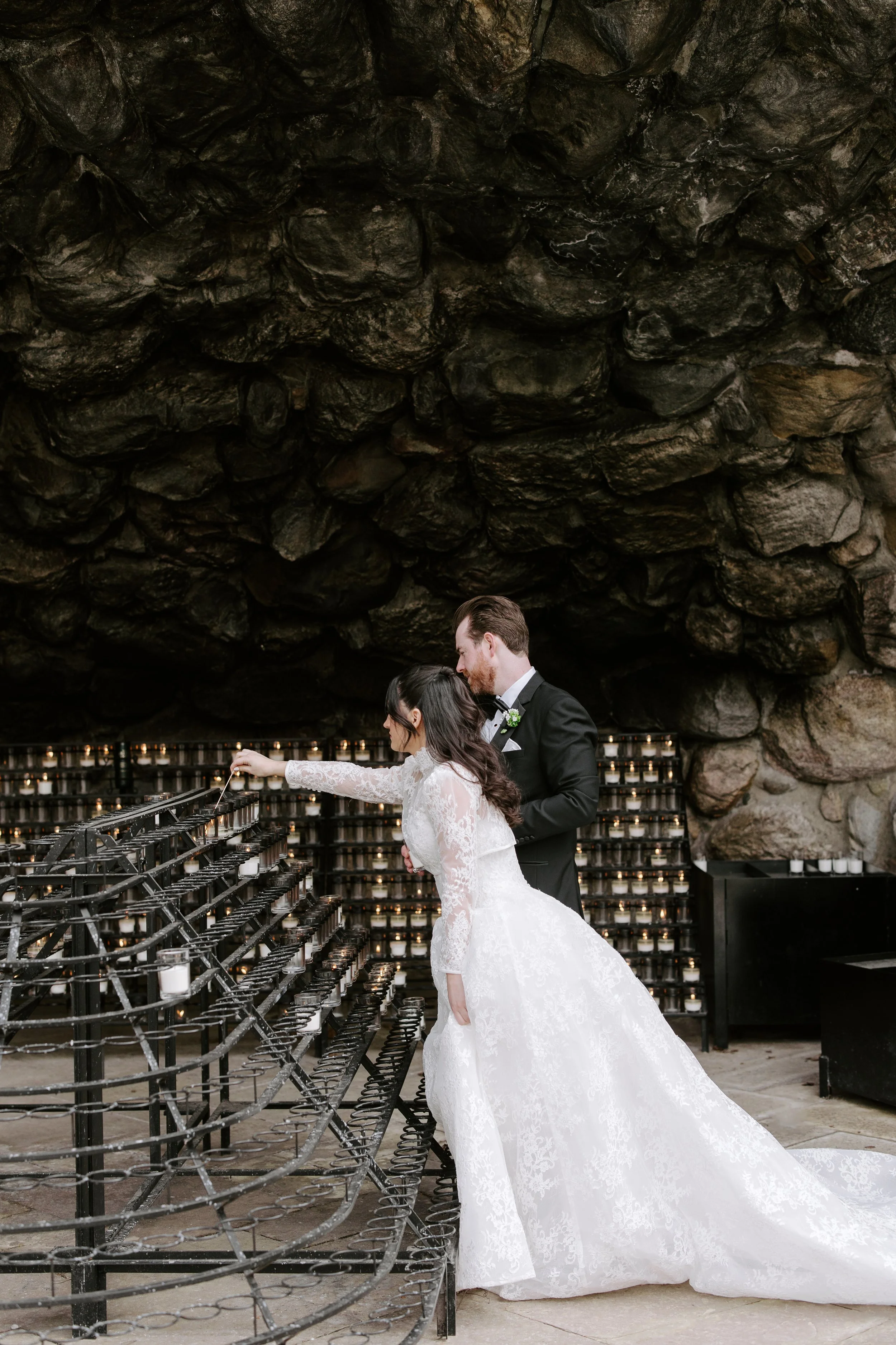 A bride and groom standing in front of a large, dark rock formation, exchanging rings during a wedding ceremony at a candlelit altar.