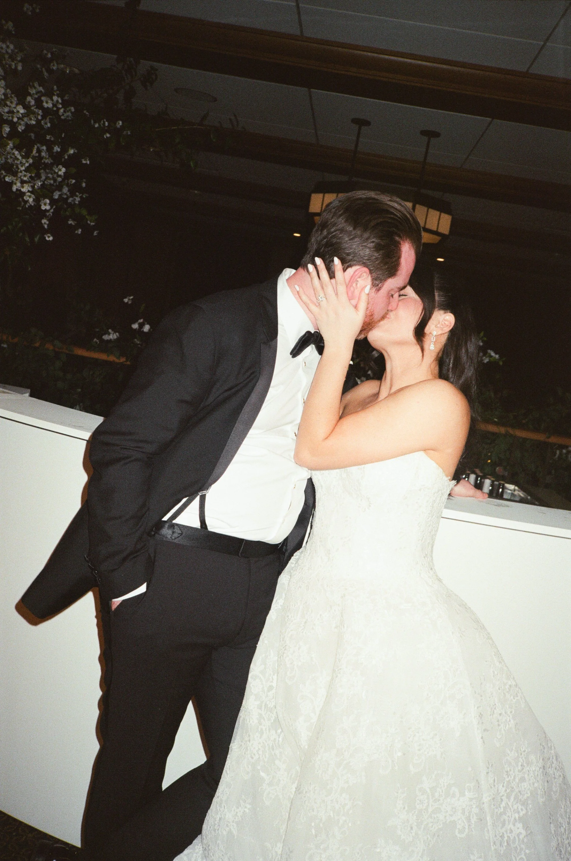 A couple in formal wedding attire kissing, with the bride wearing a lace wedding dress and the groom wearing a black tuxedo, in an indoor venue.