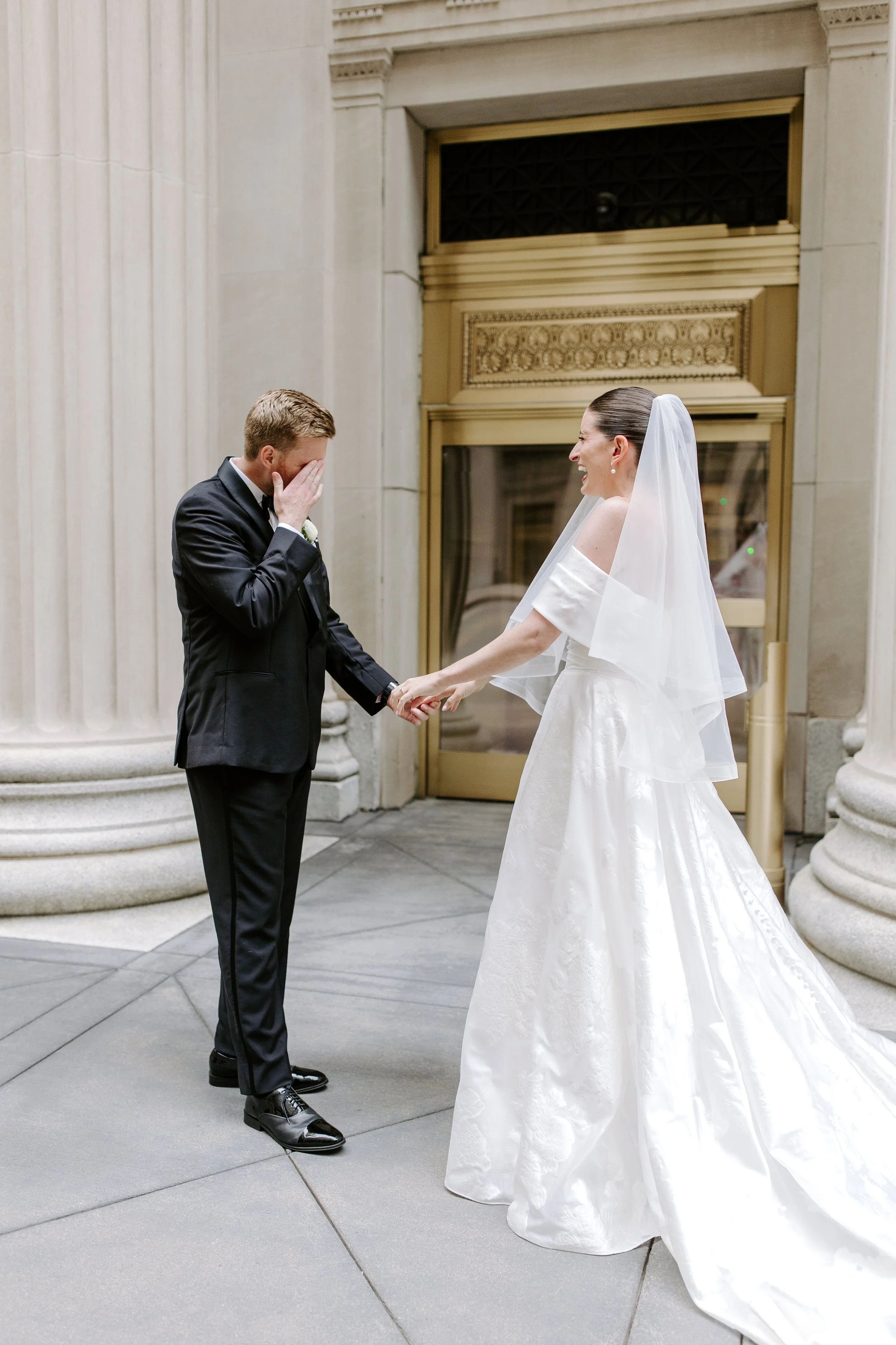 A bride in a white wedding dress and a groom in a black tuxedo are holding hands inside a building with classical architecture. The groom is covering his face with his hand, and the bride is smiling and looking at him, wearing a veil. The setting fea