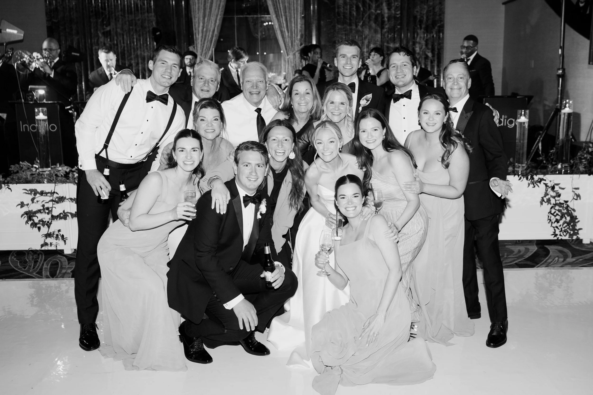Group of people dressed formally at a celebration or party, smiling for a photo, with a band playing in the background.