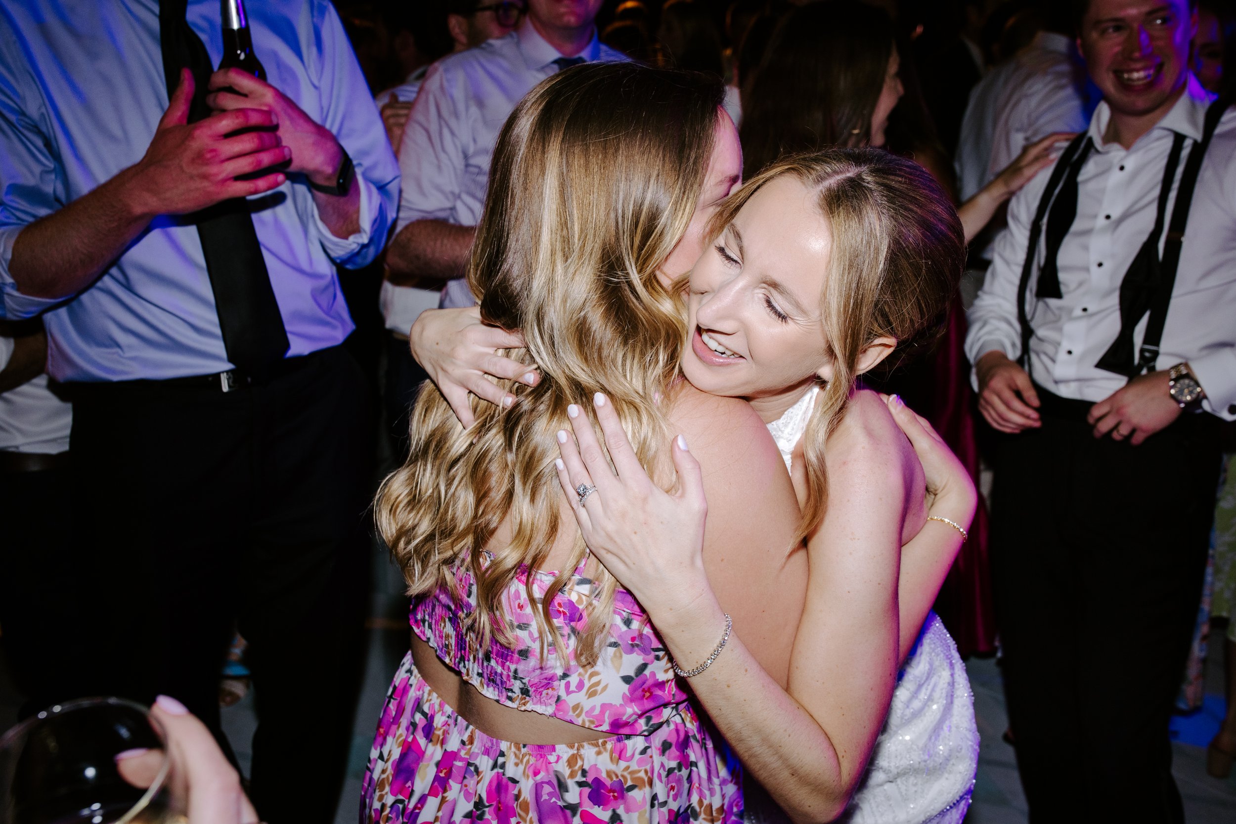 Two women hugging and smiling at a celebration or party, surrounded by other guests in formal attire.