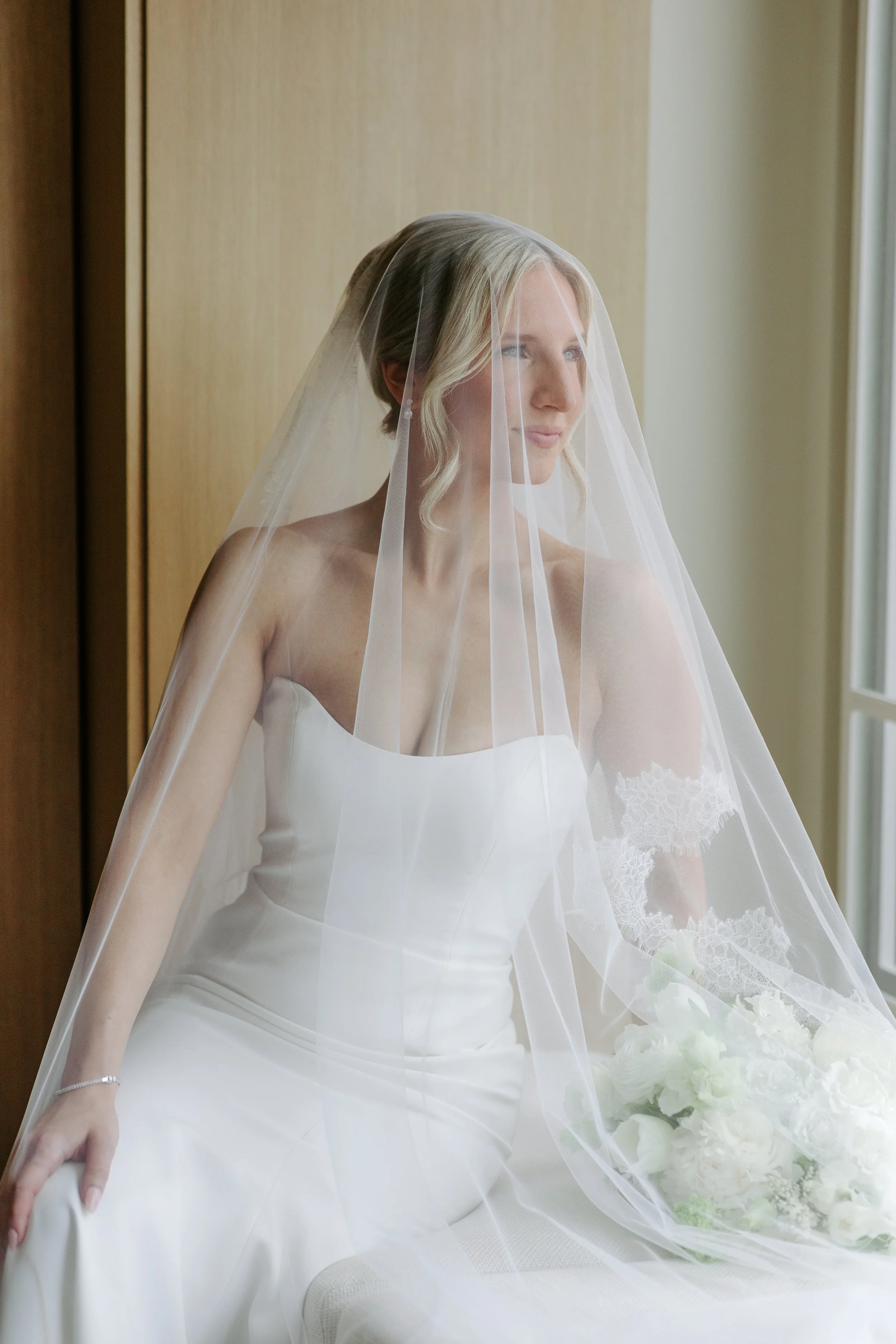 A bride in a white wedding dress with lace sleeves, sitting near a window with a veil covering her face and shoulder, holding a bouquet of white flowers.
