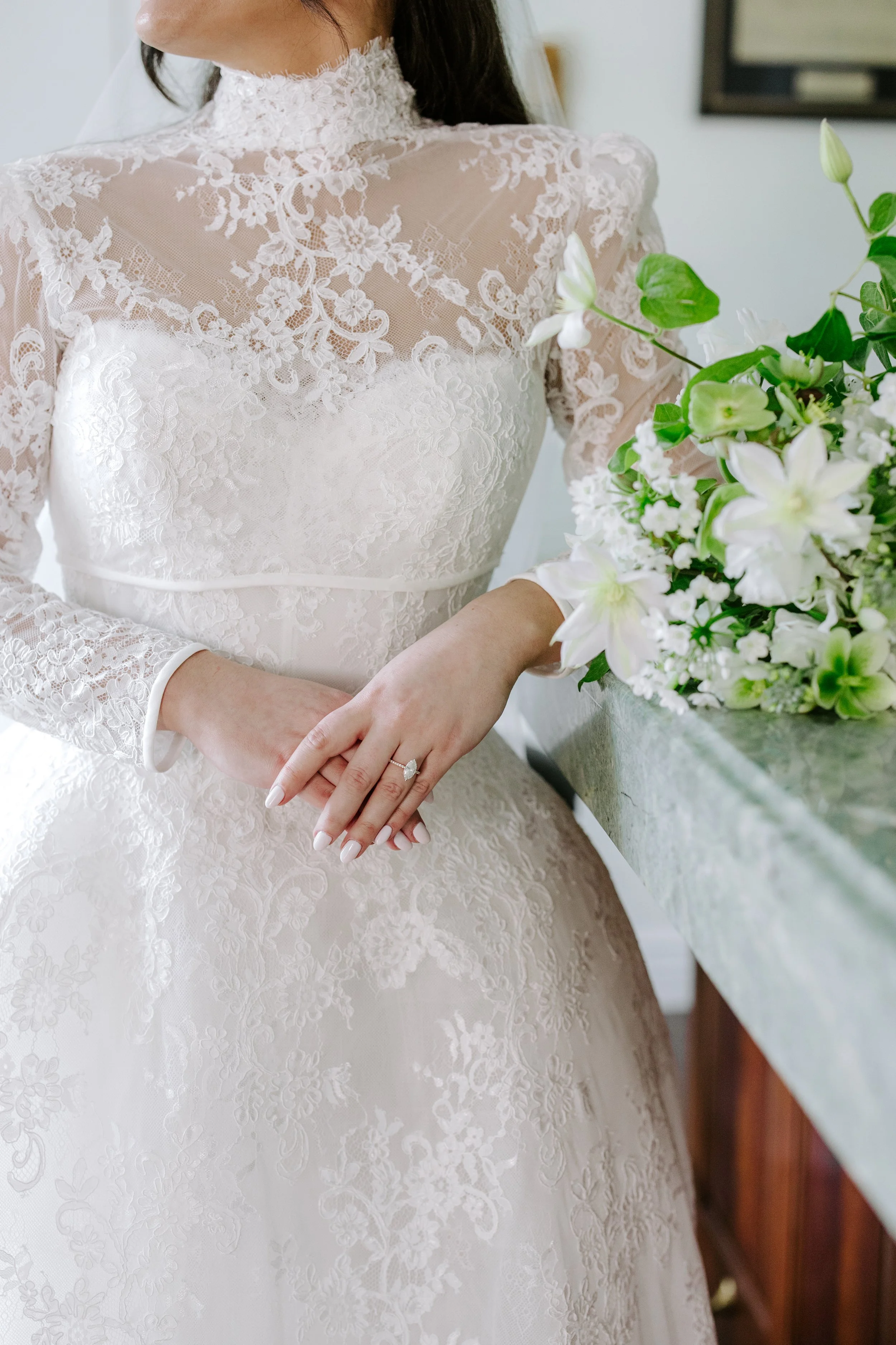 A bride in a white lace wedding dress standing near a table with a bouquet of white flowers. She is wearing a ring on her left hand.