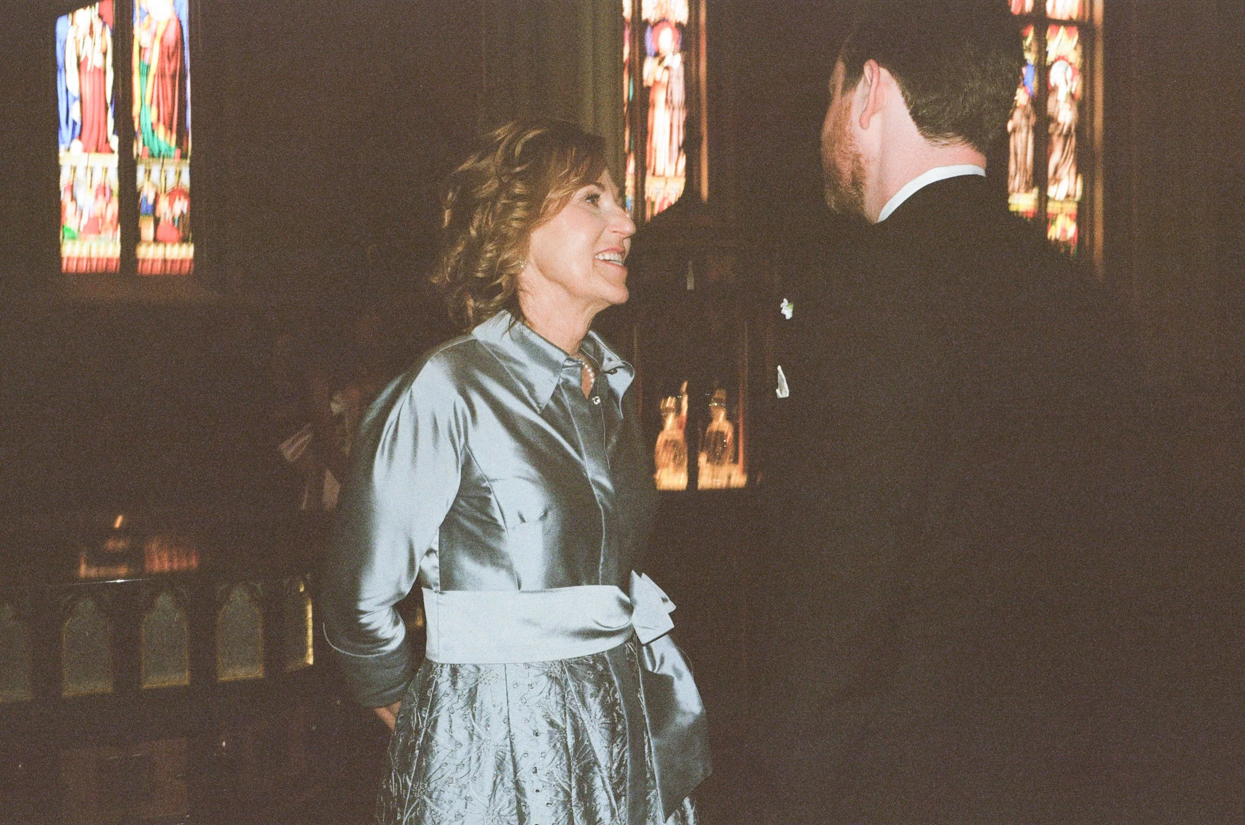 A woman and a man are standing inside a church, facing each other and smiling. The woman has curly hair and is wearing a gray satin blouse and a patterned skirt. The man has dark hair and a beard, dressed in a black suit with a white shirt. There are