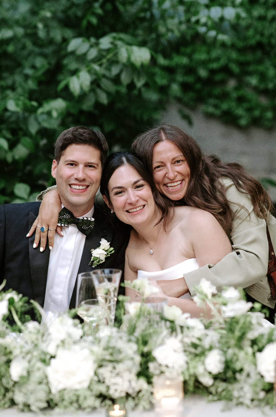 Three people smiling and hugging at a wedding reception, with floral decorations on the table in the foreground and a green leafy background.