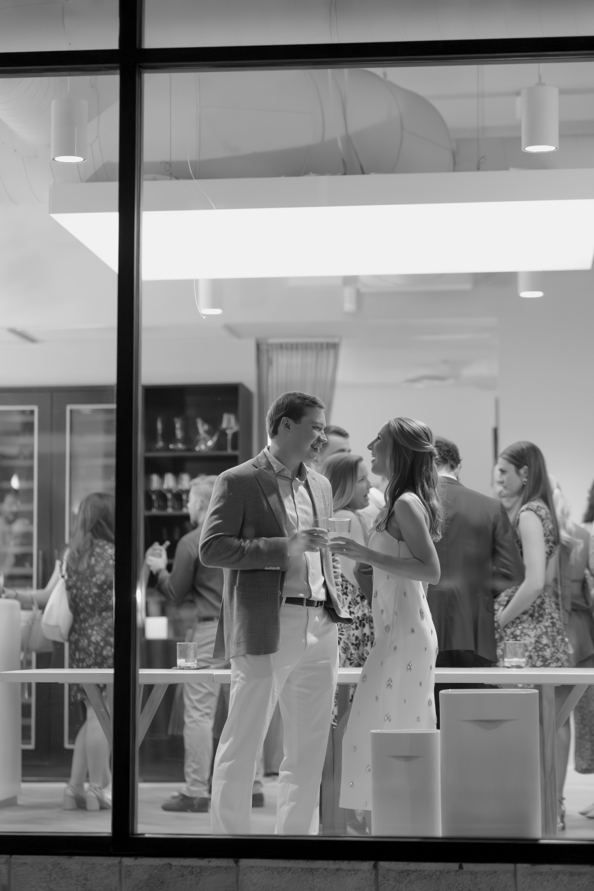 People socializing and chatting at a modern indoor event, viewed through a glass window, in black and white.