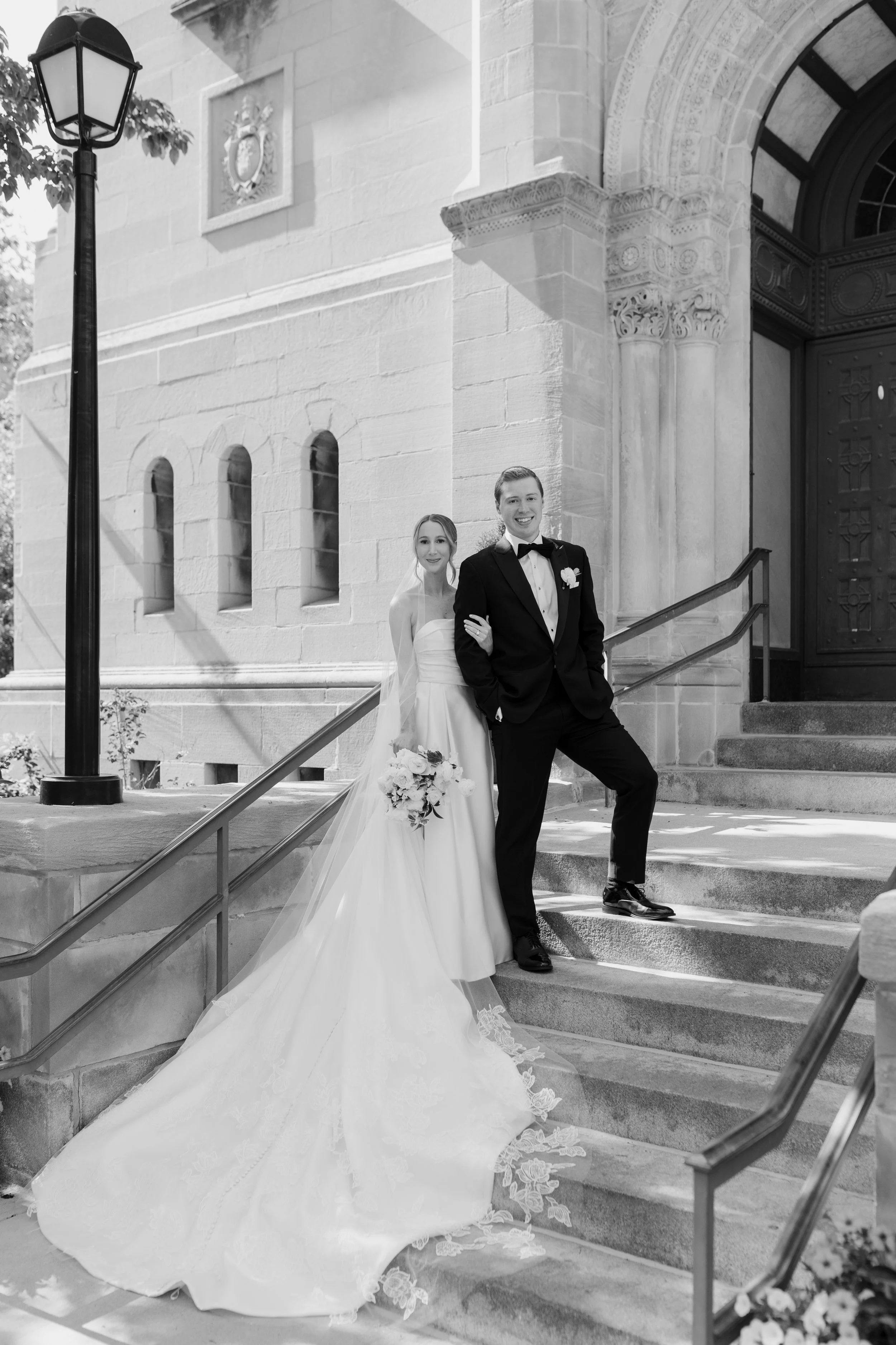 A newlywed couple poses on the steps outside a stone church. The bride is in a long white wedding gown with a train and holds a bouquet of flowers. The groom is in a black tuxedo with a bow tie, standing with one hand in his pocket, smiling at the ca