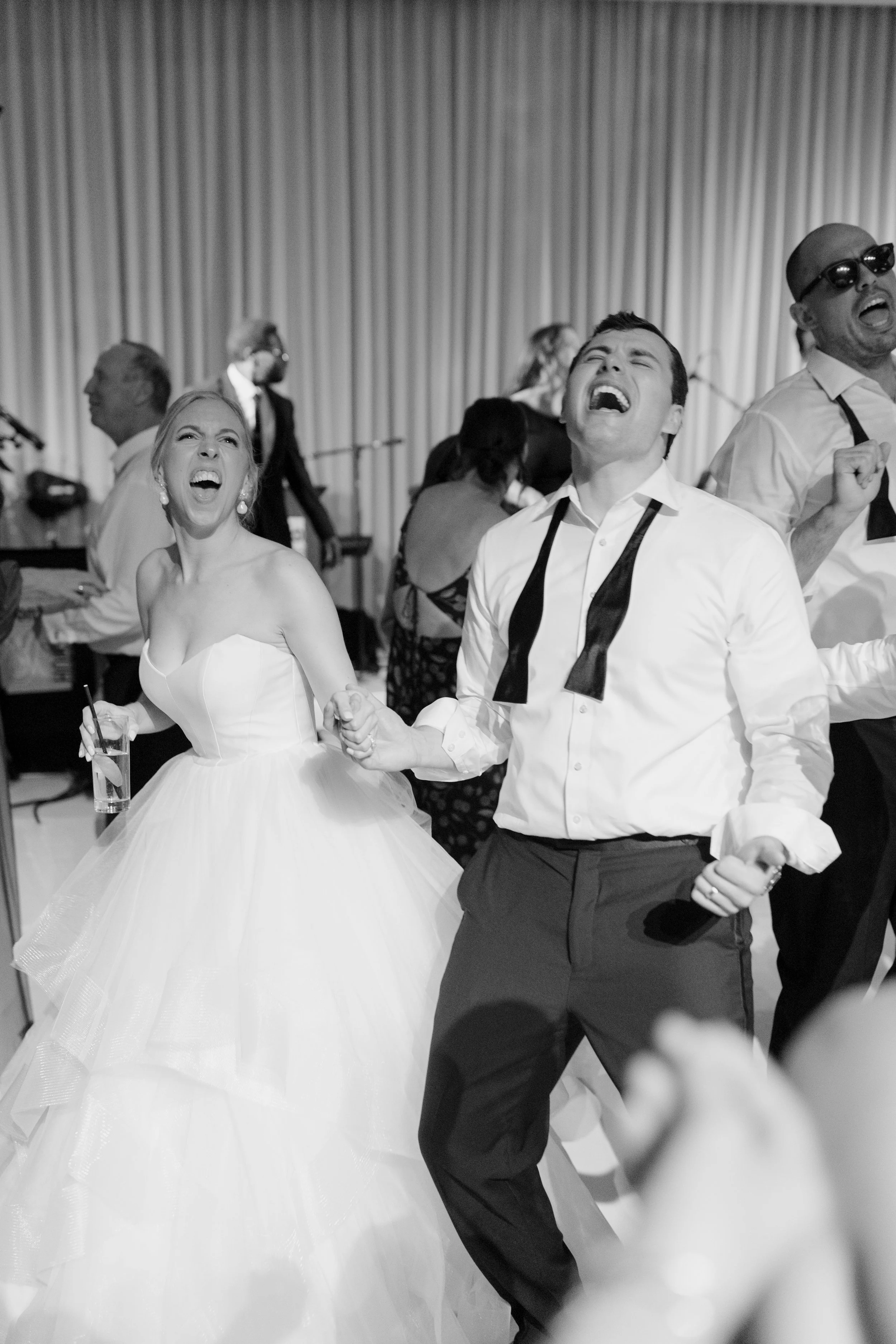 A black and white photo of a bride and groom dancing and singing at their wedding reception, with joyful expressions. The bride is wearing a strapless wedding dress and the groom is wearing a white shirt and dark dress pants.