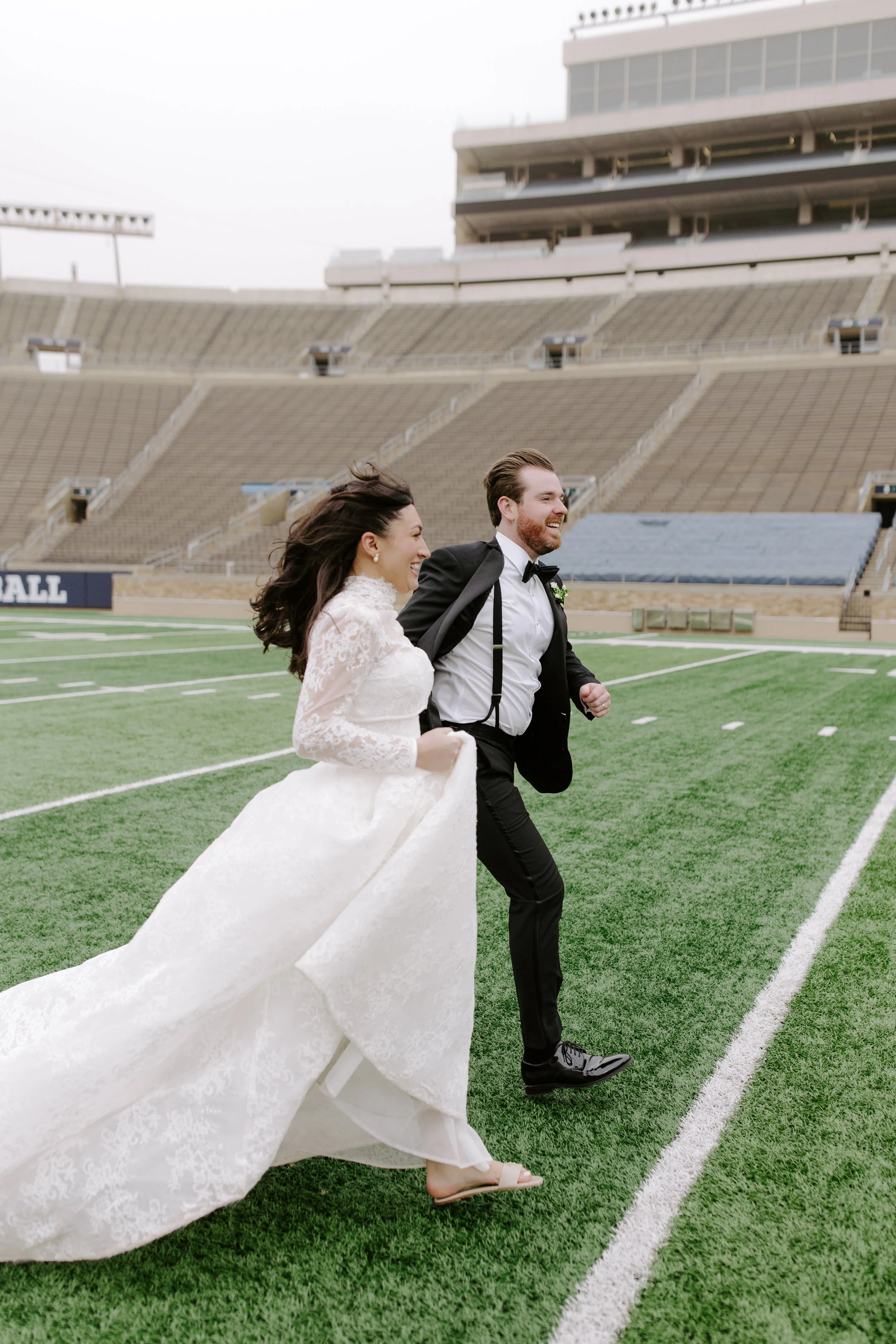 A bride and groom running on a football field in a stadium. The bride is wearing a white lace wedding dress, and the groom is in a black tuxedo with suspenders and a bow tie.