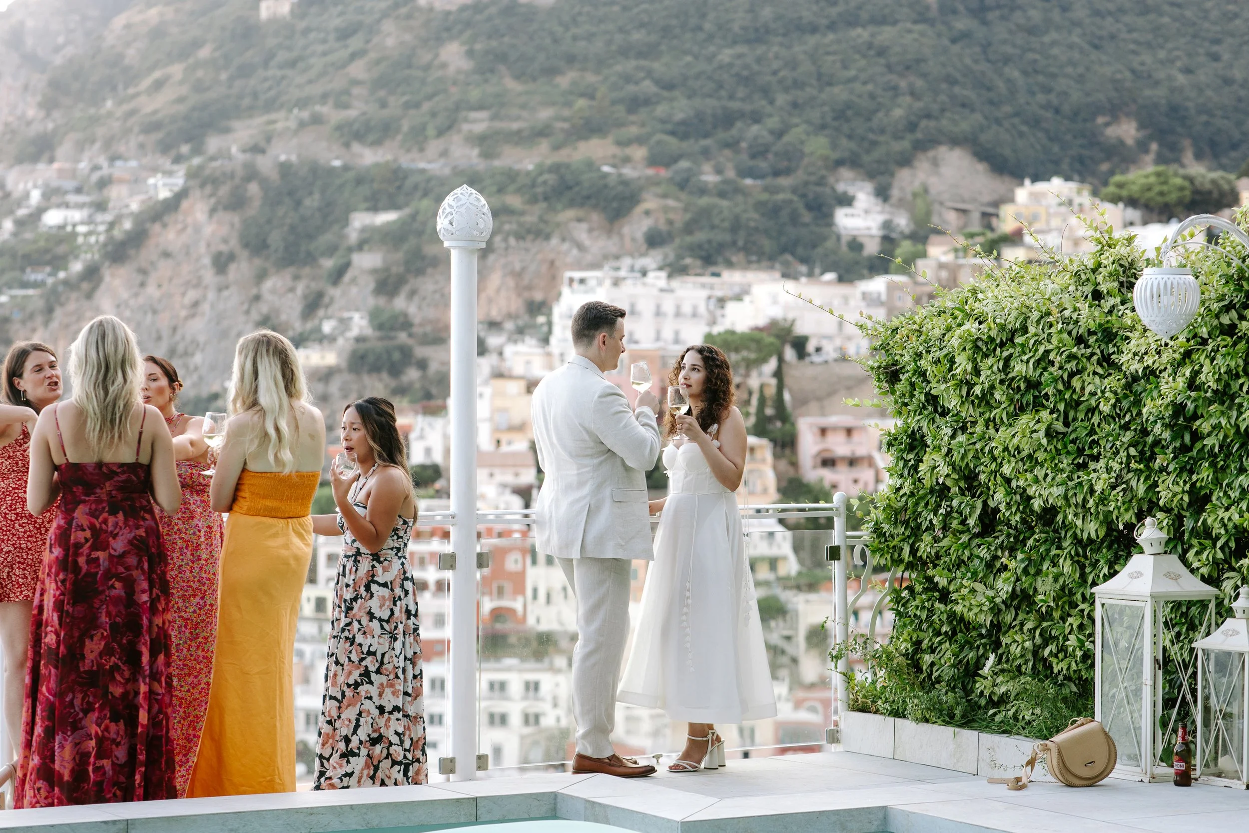 A group of people, mostly women in colorful dresses, at an outdoor gathering, with a man and woman in the center holding wine glasses, against a hillside with buildings.
