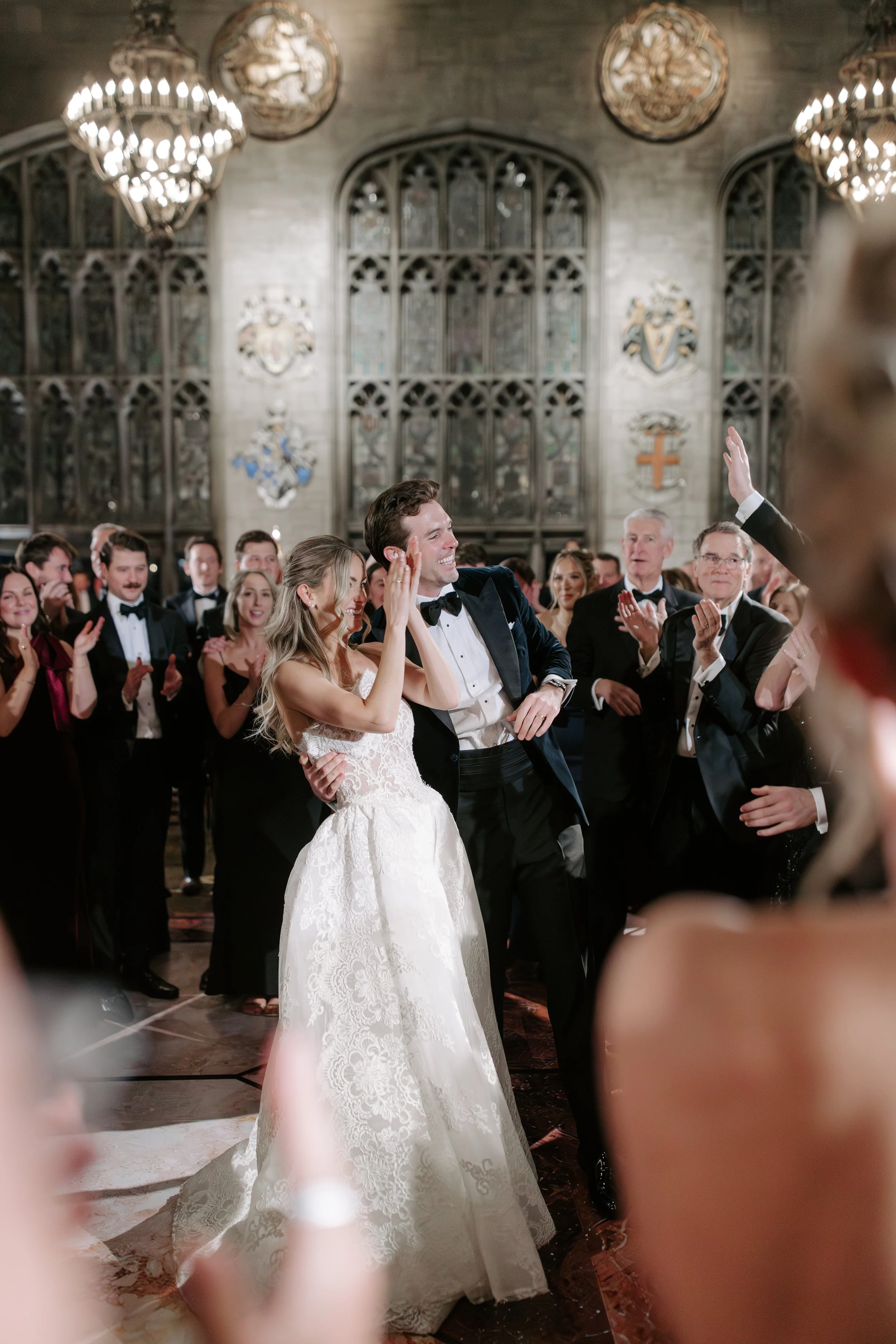 A bride and groom dancing at their wedding reception, surrounded by guests in a church or cathedral with stained glass windows and coat of arms on the walls.