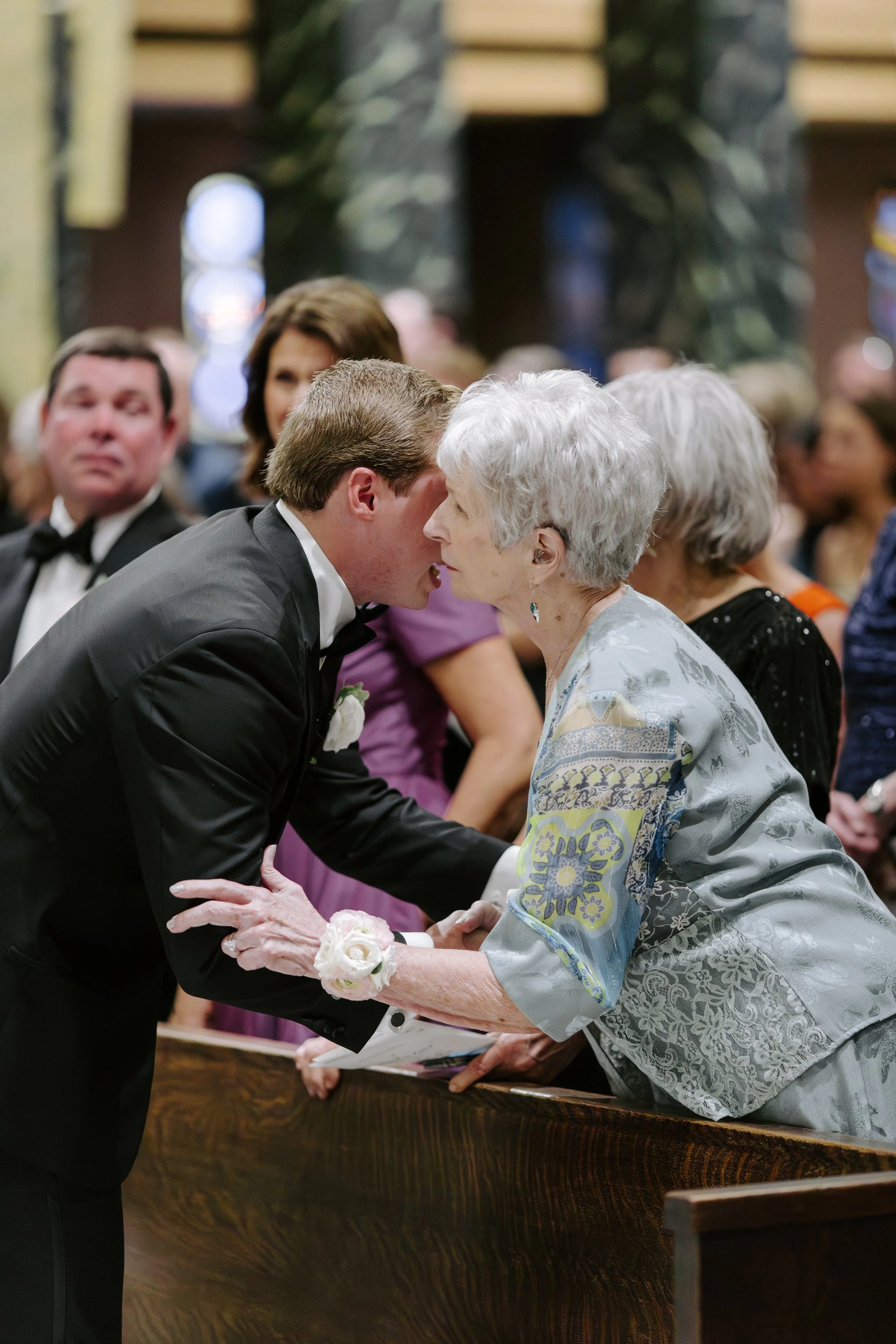 'A young man in a tuxedo gently holding hands with an elderly woman dressed in a floral jacket, both with closed eyes, moment of tender interaction at a formal gathering, with other attendees blurred in background.