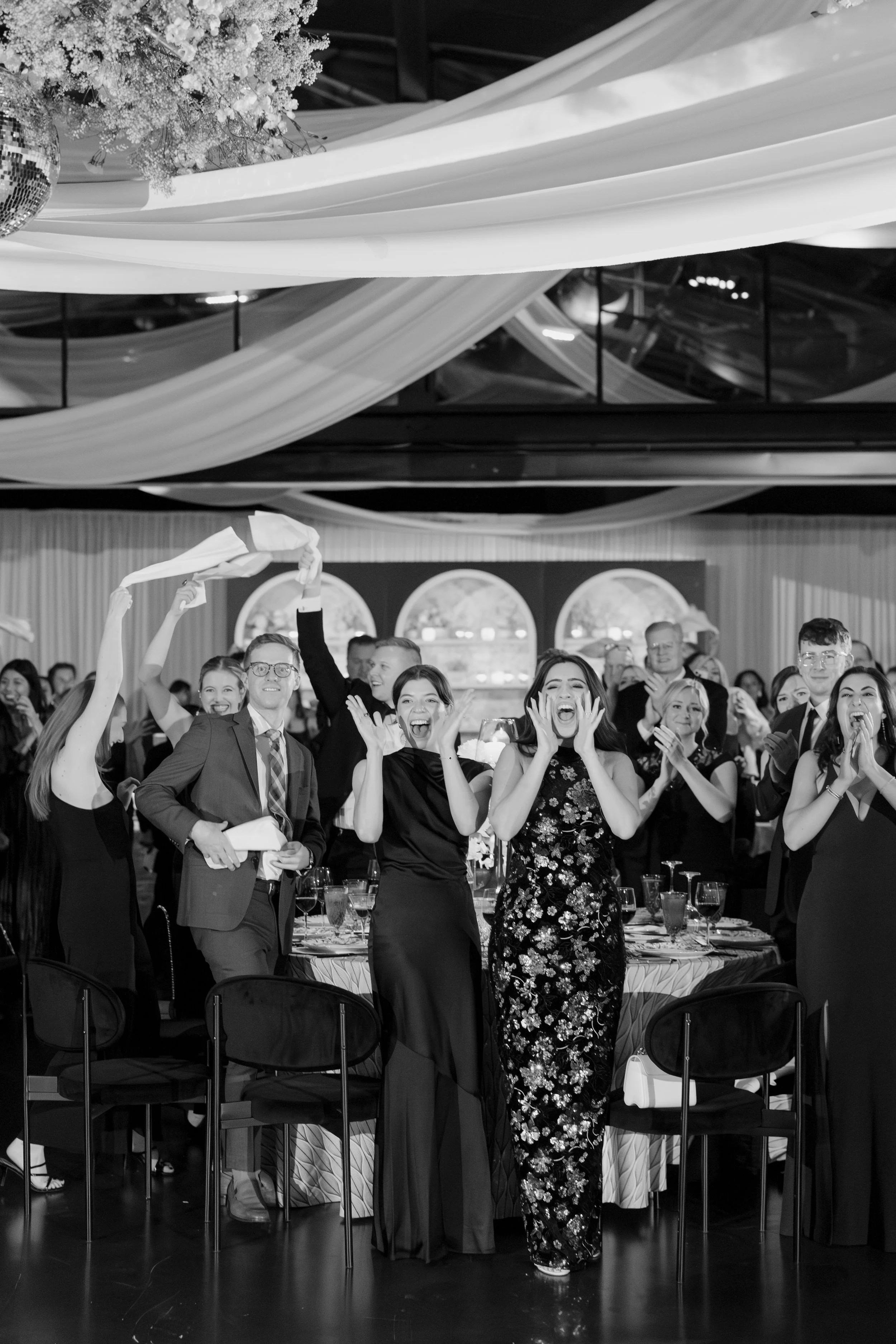 People at a joyful celebration or party, dressed formally, with some raising their hands and smiling, in a decorated venue with drapes and a mirror ball, set for a banquet or reception.