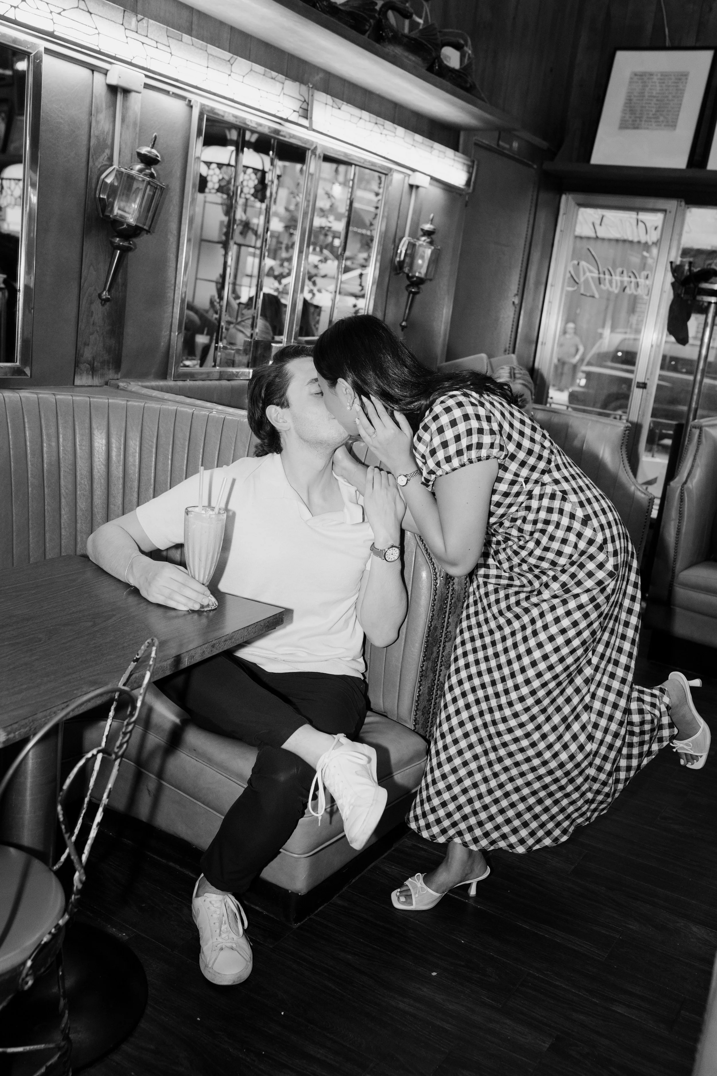 A woman in a checkered dress is kneeling on the floor and kissing a man who is sitting on a booth seat in a diner. The man has a drink with a straw on the table in front of him.