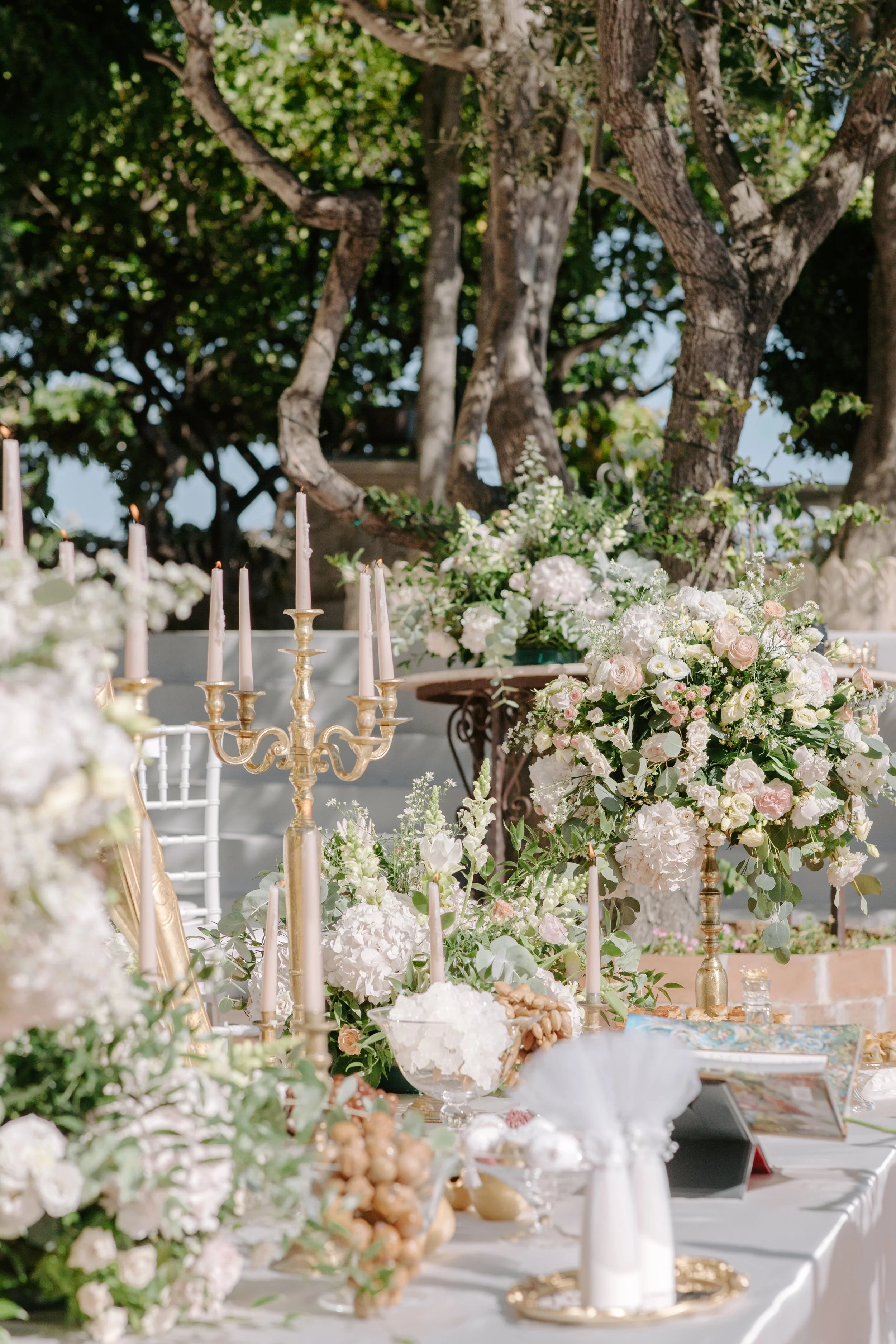 A beautifully decorated outdoor table with white and pink floral arrangements, gold candlesticks with pink candles, and elegant tableware, set against a backdrop of lush trees and sunlight.