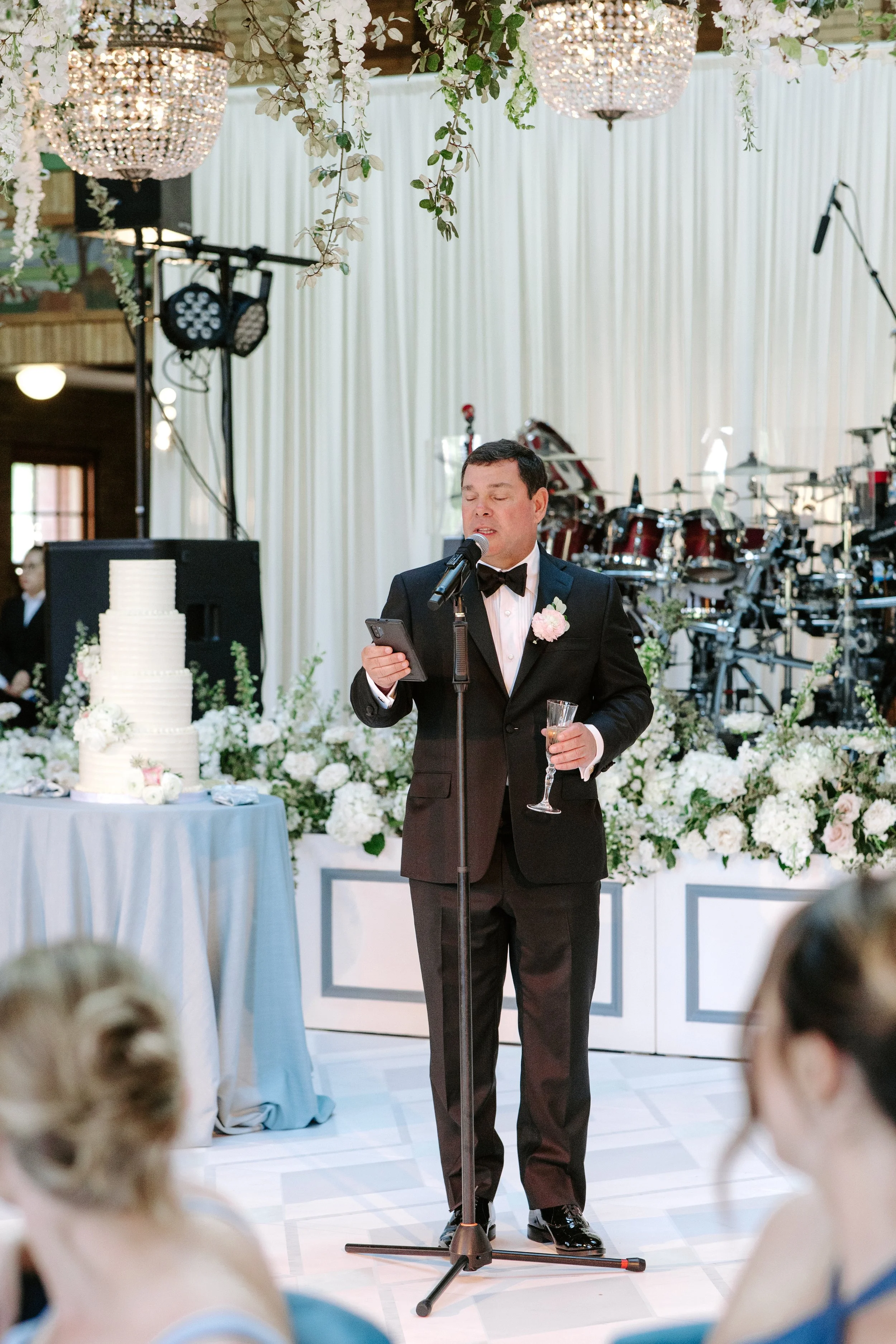A man in a tuxedo with a pink boutonniere is speaking into a microphone while holding a glass of champagne at a wedding reception, with a decorated wedding cake and floral arrangements in the background.