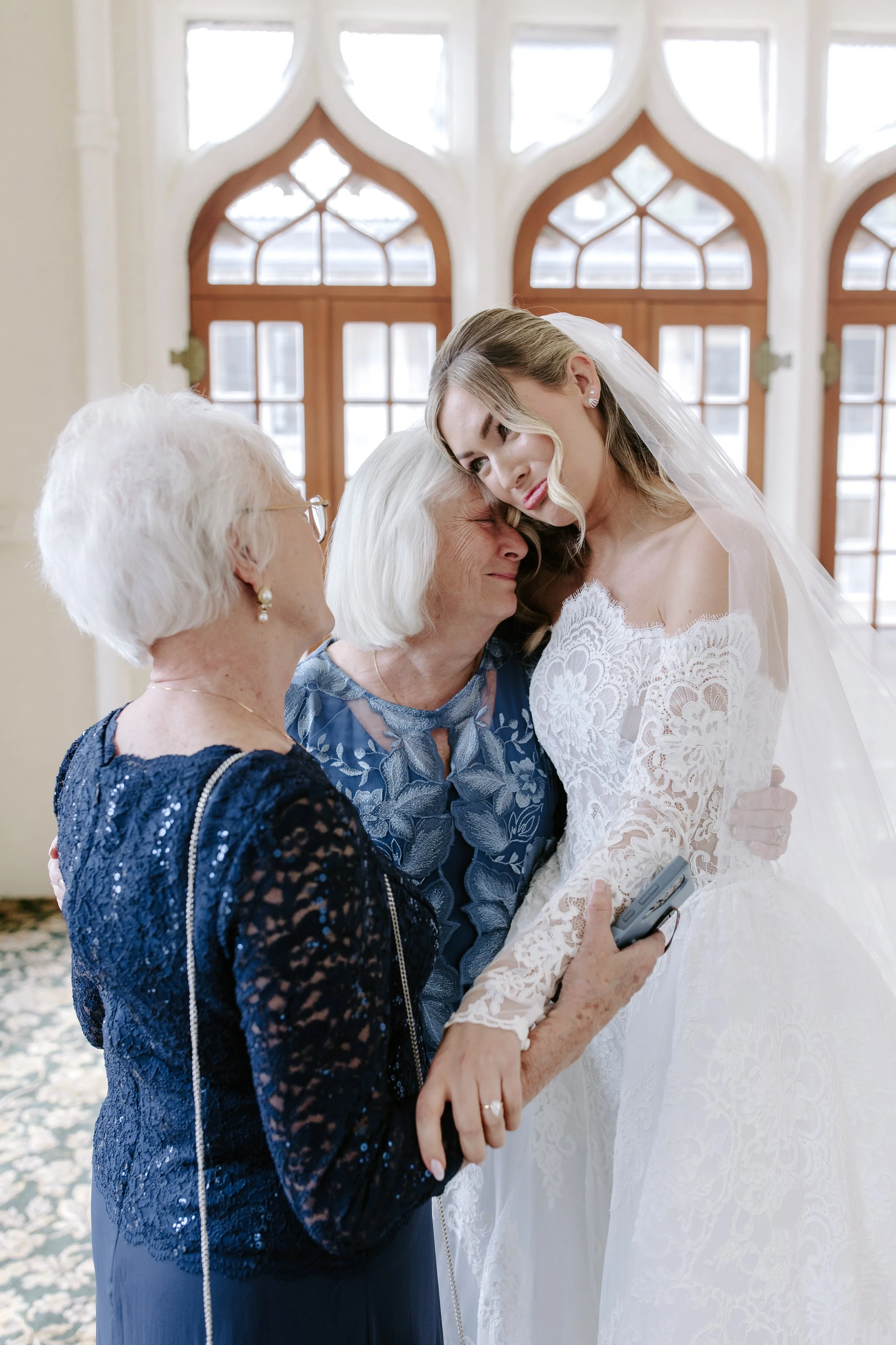 A bride in a white lace wedding dress and veil is hugging her elderly grandmother, who has short white hair and glasses, and is wearing a dark blue sequined dress, inside a bright room with arched wooden windows.