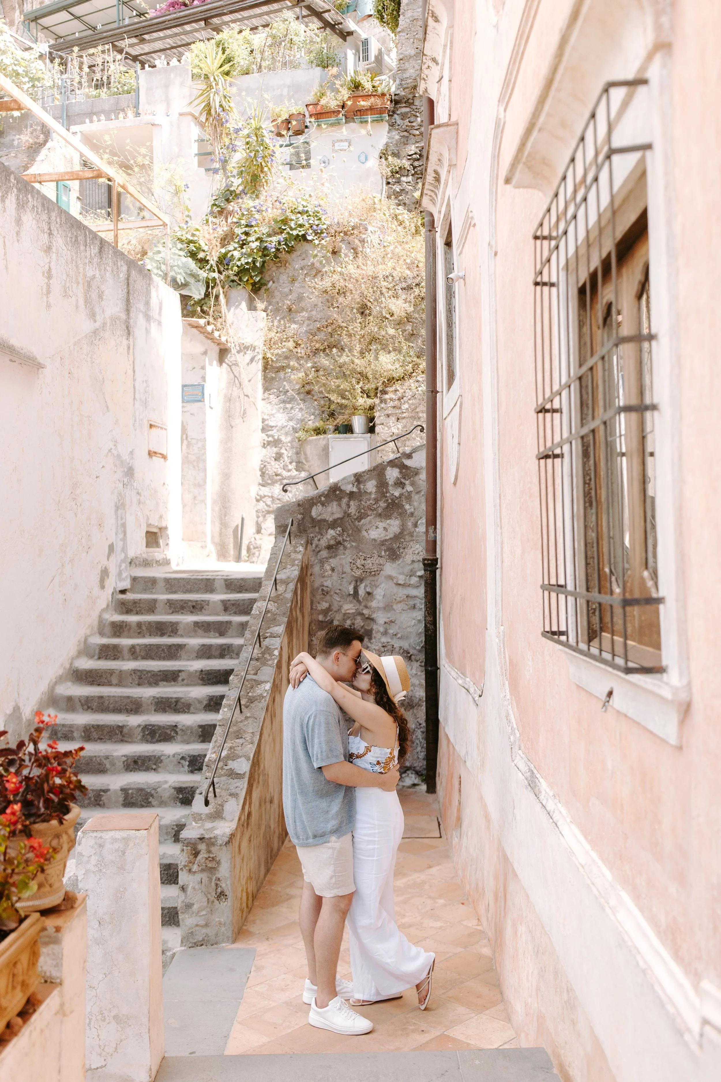 A couple kissing in a narrow, sunny alley with stone stairs and old walls, with potted plants and flowers.