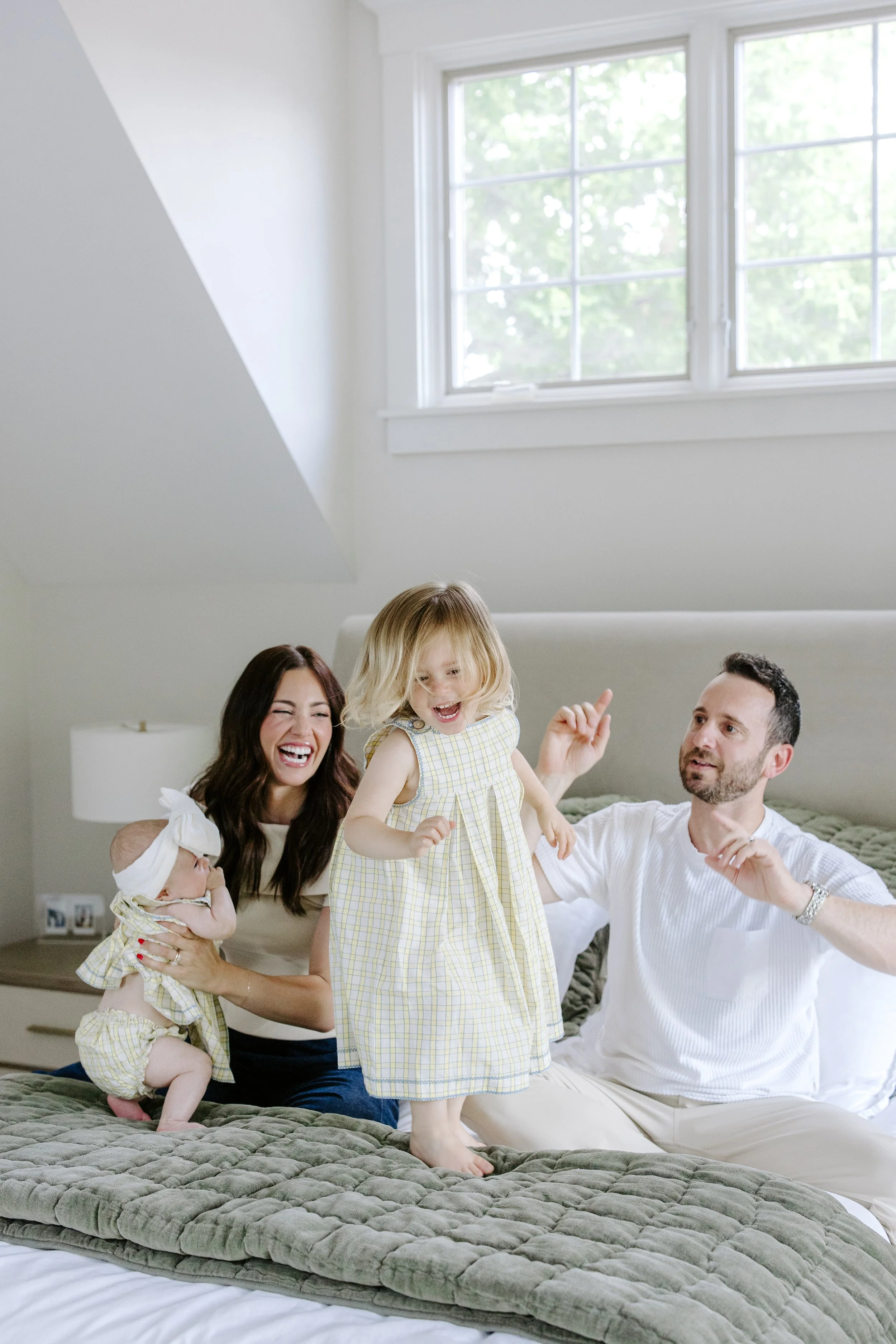 A family of four on a bed in a bright bedroom with a large window. The mother and father are smiling and holding their young daughters, who are dressed in matching yellow checkered dresses. The family appears happy and playful.