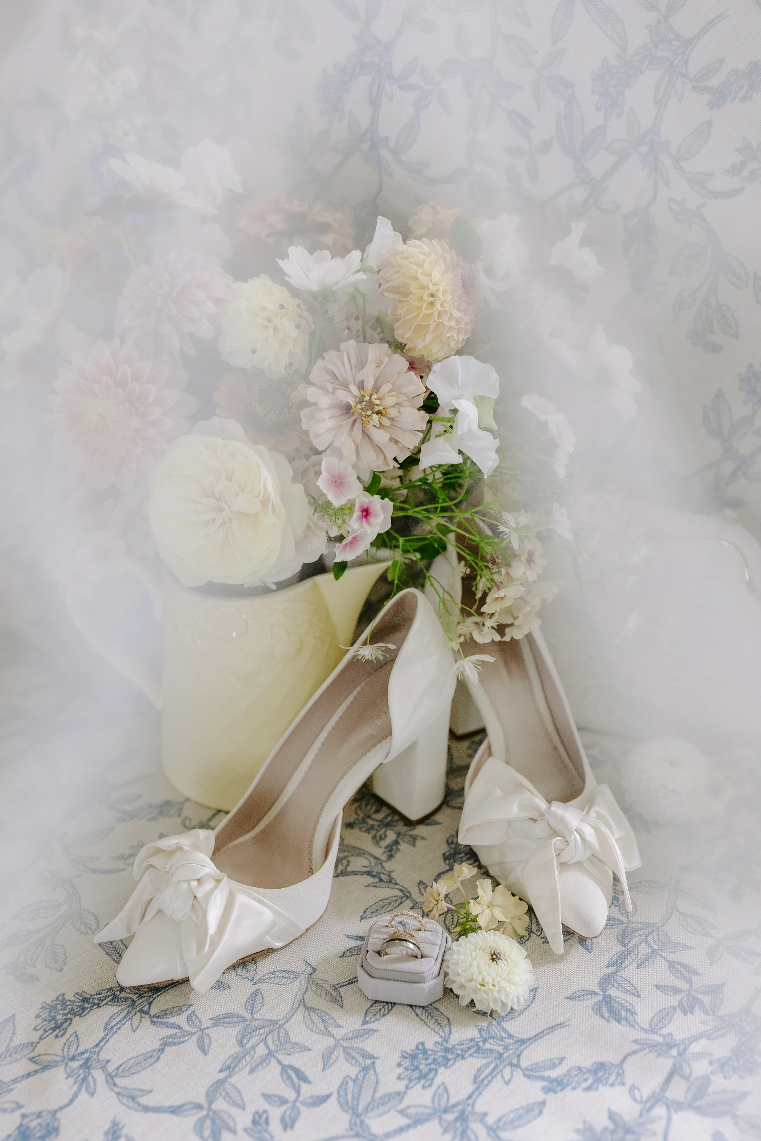 A bridal bouquet with white, cream, and blush pink flowers. A pair of white satin wedding shoes with bows and a wedding ring box with rings are also displayed, all arranged on a floral-patterned fabric background.