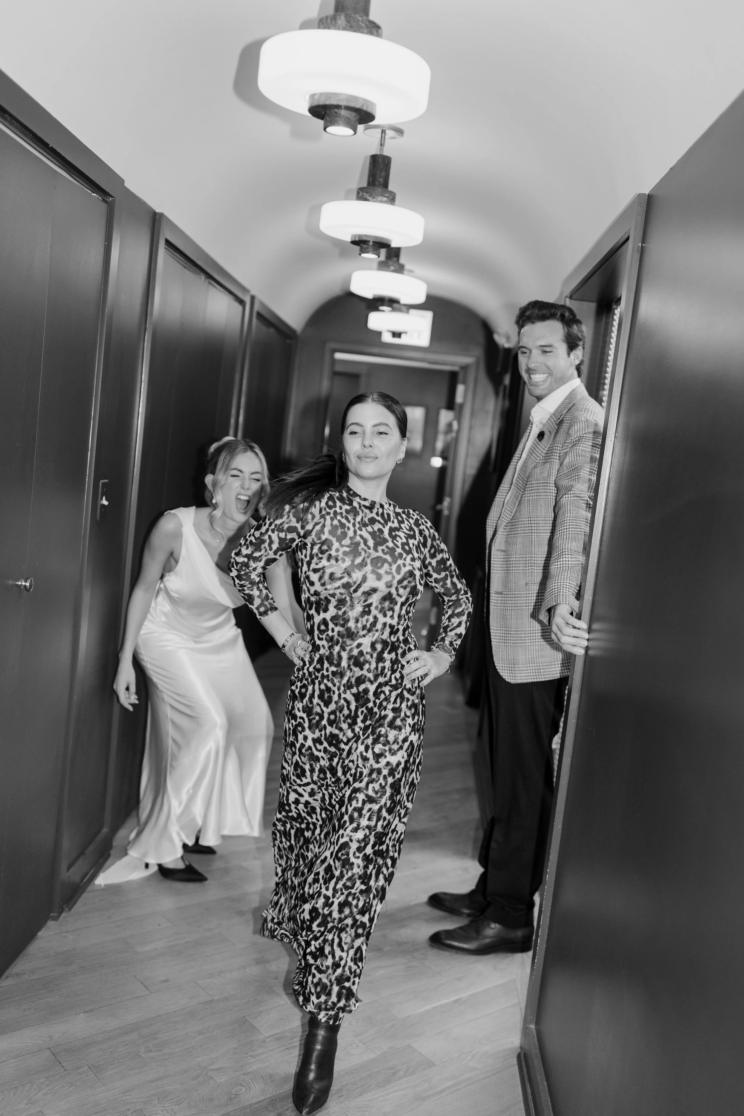 People in stylish clothing laughing and posing in a hallway with modern light fixtures and wooden flooring.