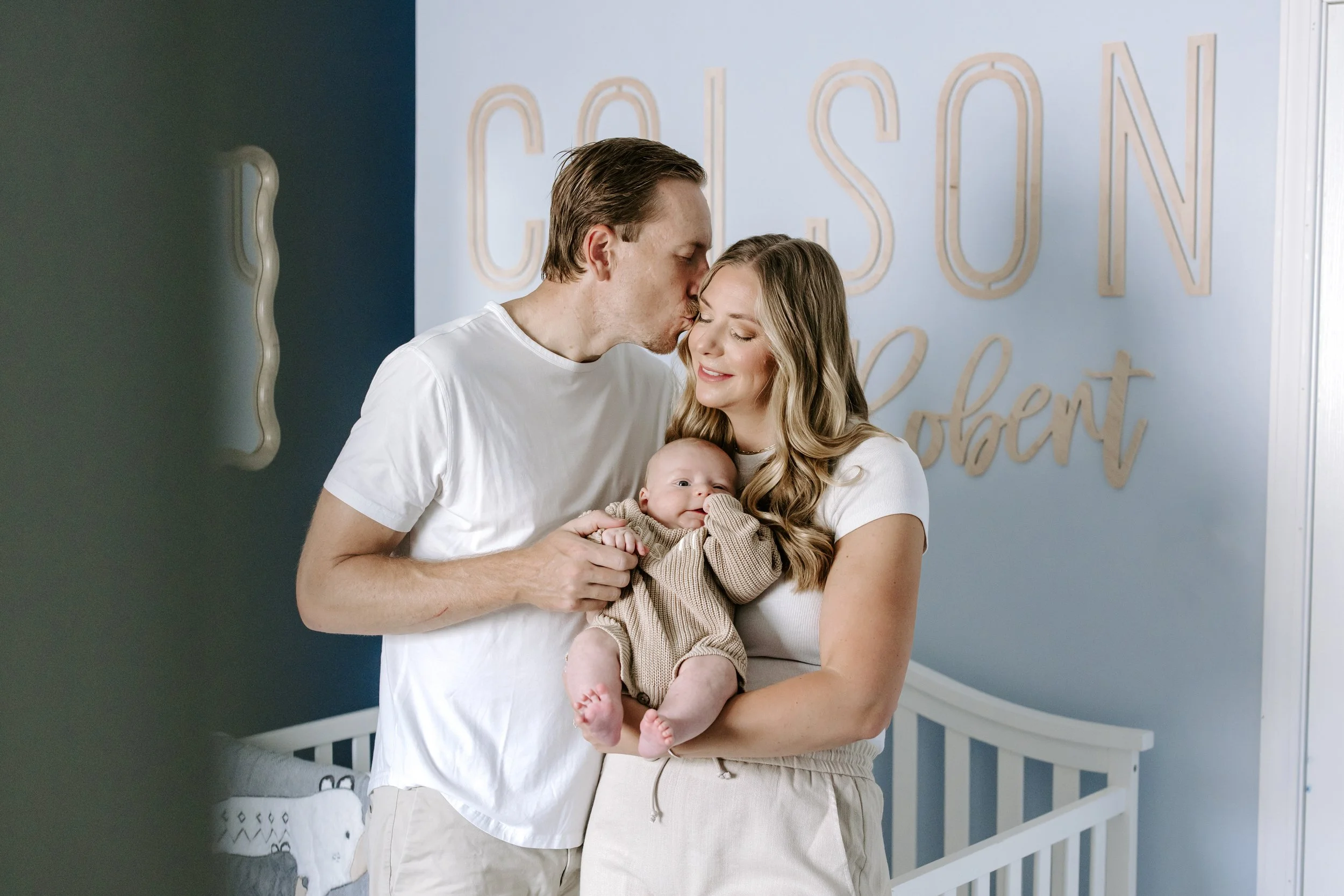 A family of three standing in a nursery room, with a dark blue wall on the left and a light blue wall with the words 'CASON' and 'present' in decorative letters on the right. The man, woman, and baby are smiling and hugging, with the man kissing the 