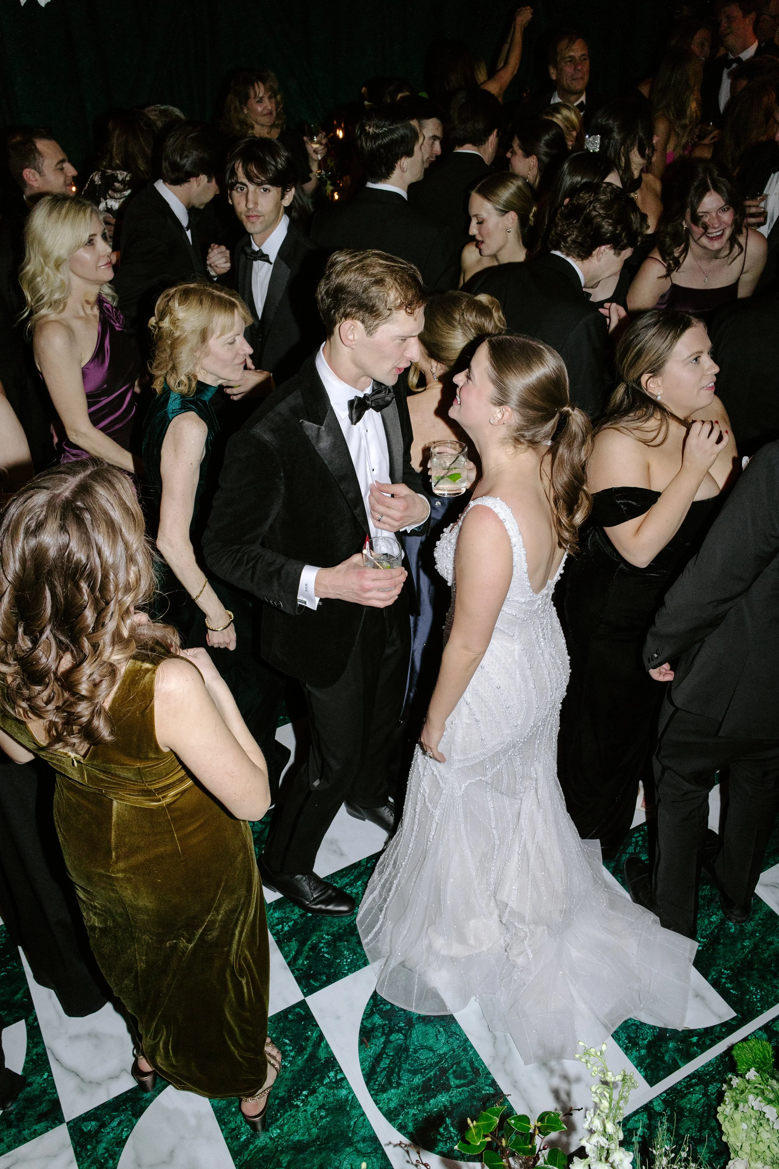 A wedding reception with many guests, a bride in a white gown, and a groom in a tuxedo, standing on a black and white marble patterned floor, with people socializing and holding drinks.