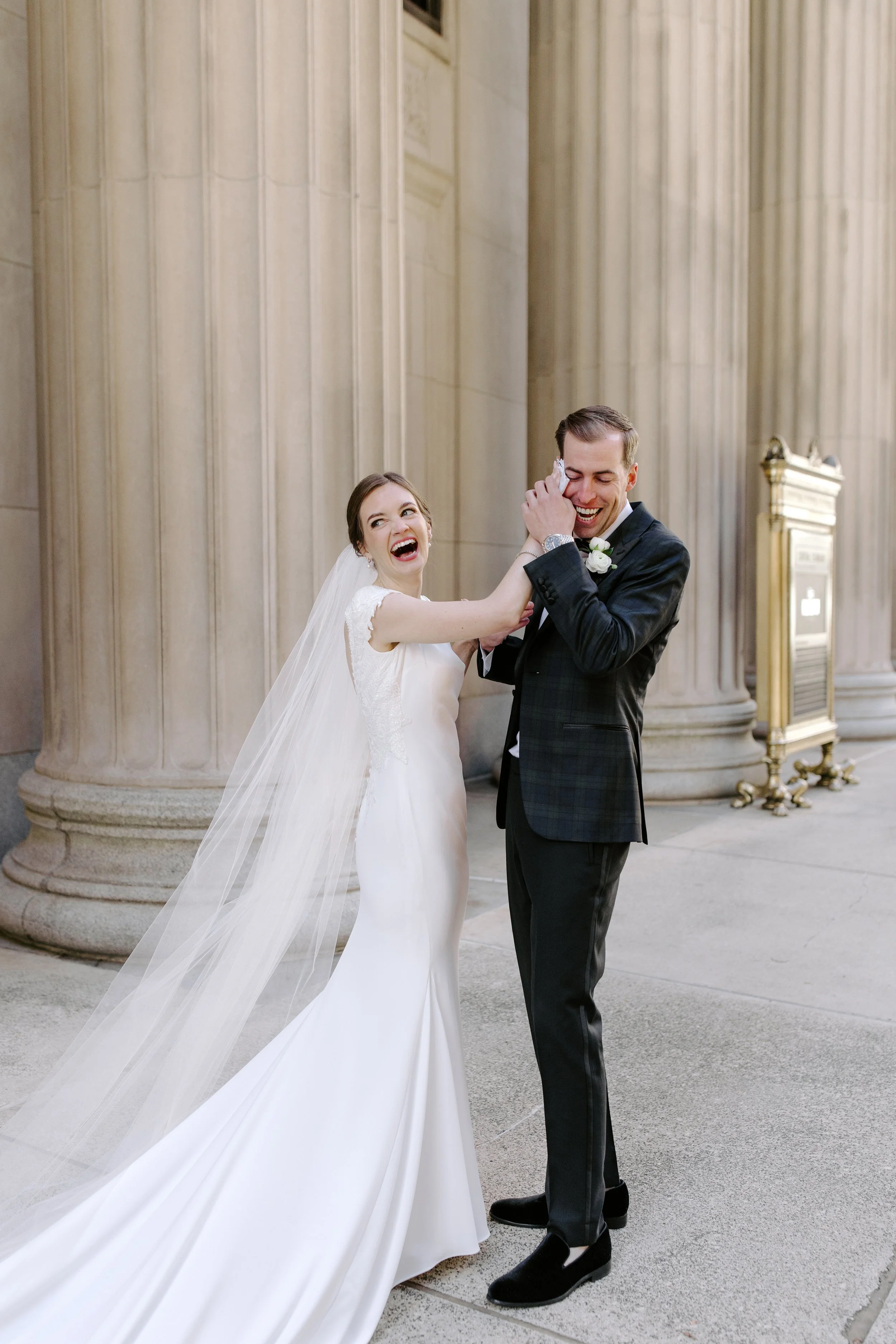 A bride and groom are laughing and having fun outside a building with large stone columns. The bride is wearing a white wedding dress and veil, and the groom is in a dark suit, adjusting his tuxedo cufflink, with a white boutonniere on his lapel.