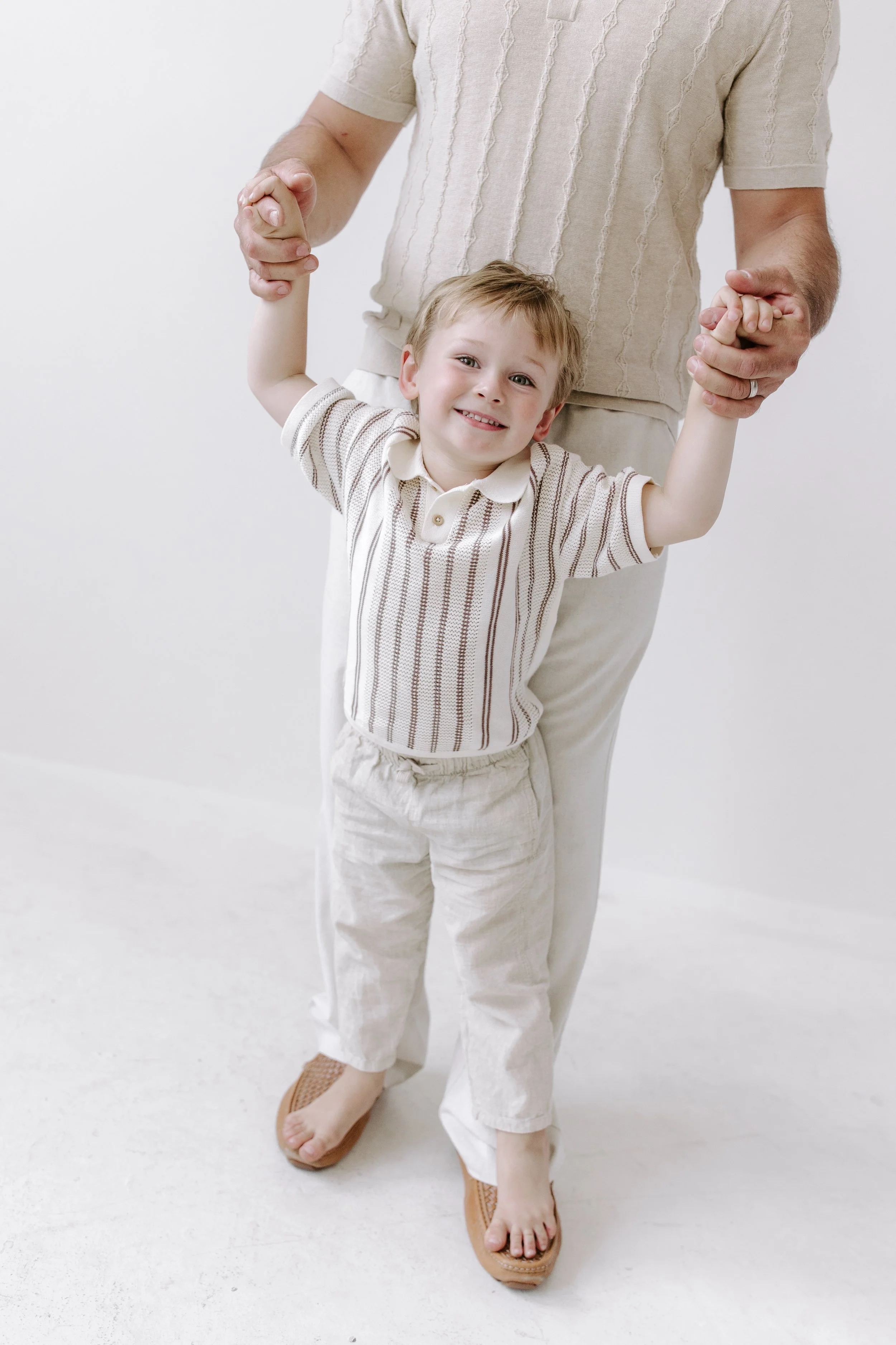 A smiling young boy standing on a white floor, holding hands with an adult whose face is not visible. The boy is wearing beige and light brown striped shirt and beige pants, with tan slip-on shoes. The adult is dressed in a beige cardigan and matchin