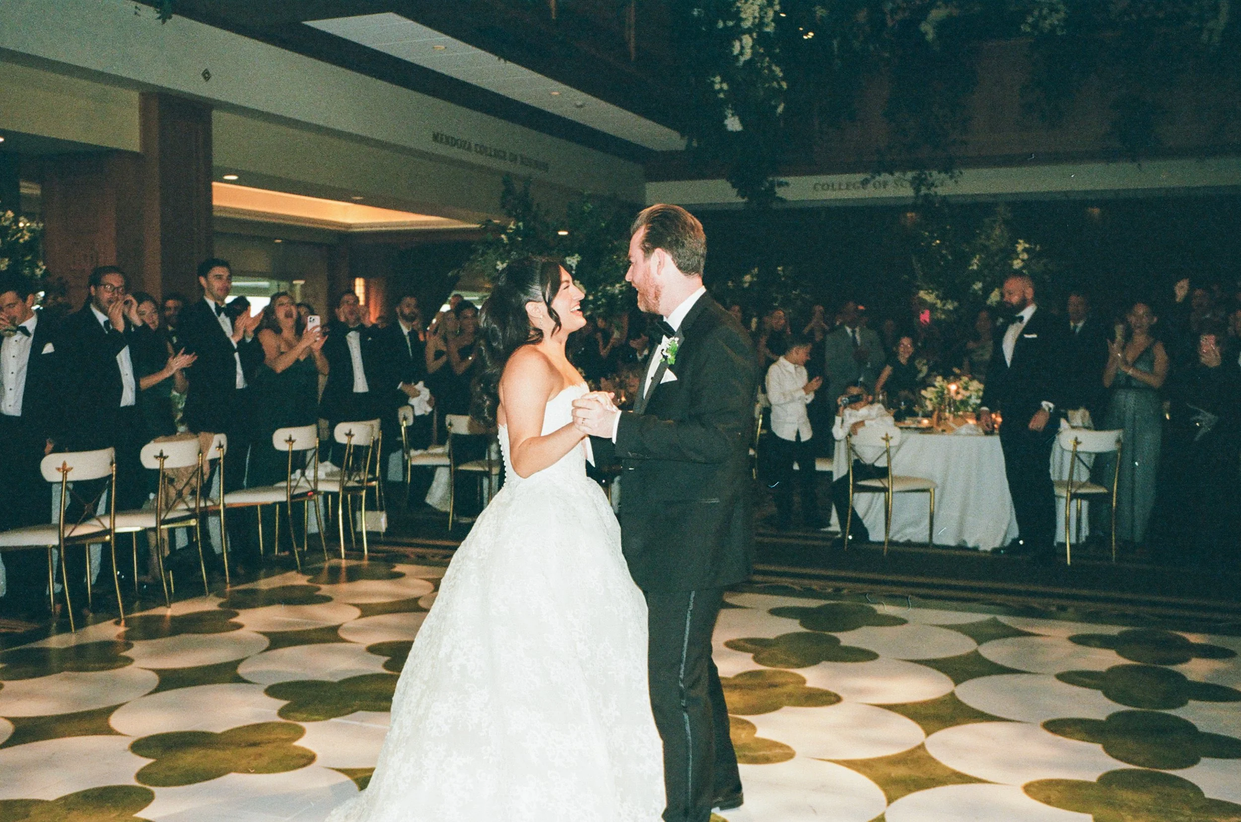 A bride and groom dance together at their wedding reception in a banquet hall, with guests clapping and smiling in the background.