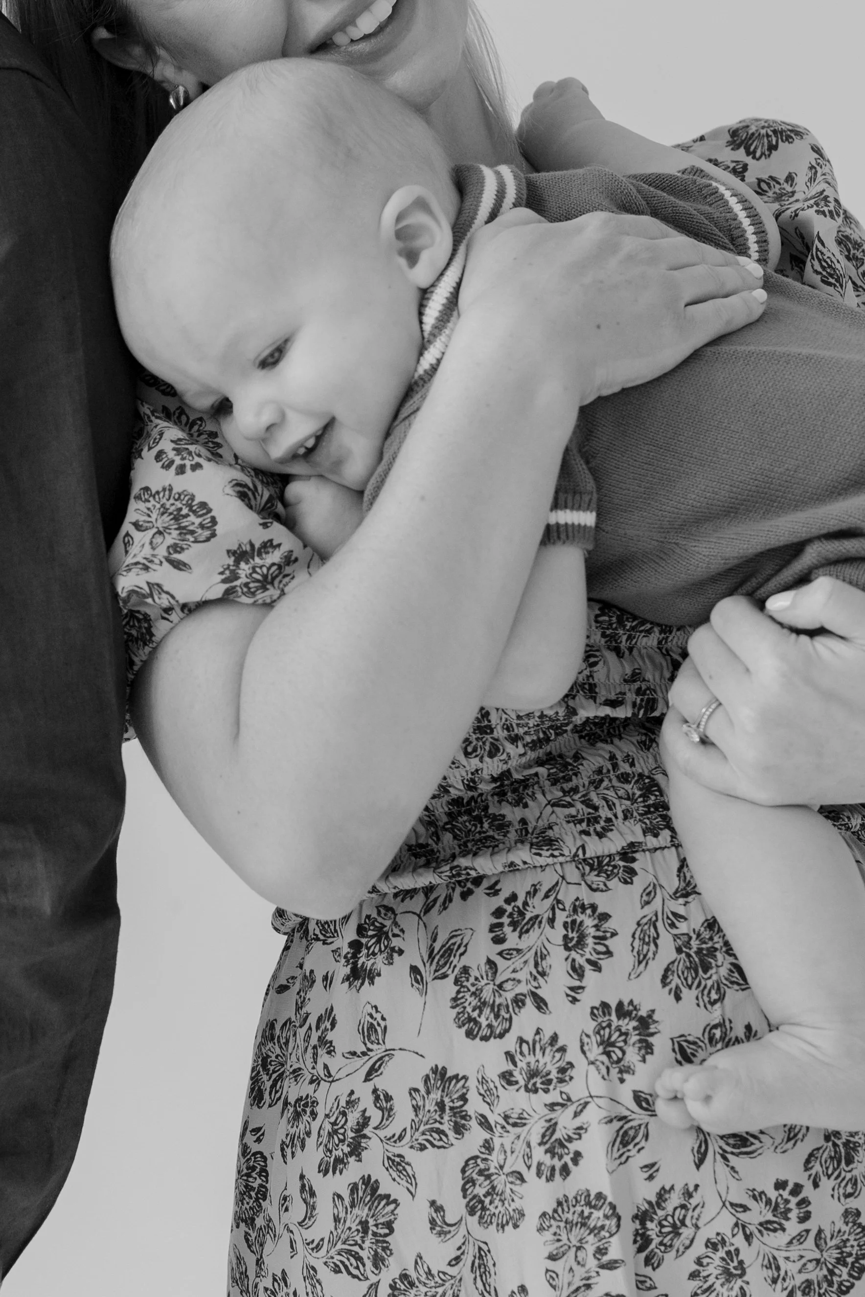 A woman holding a smiling baby in her arms. The woman is wearing a floral dress and has a ring on her finger. The baby is wearing a striped shirt and is resting their head on the woman's shoulder.