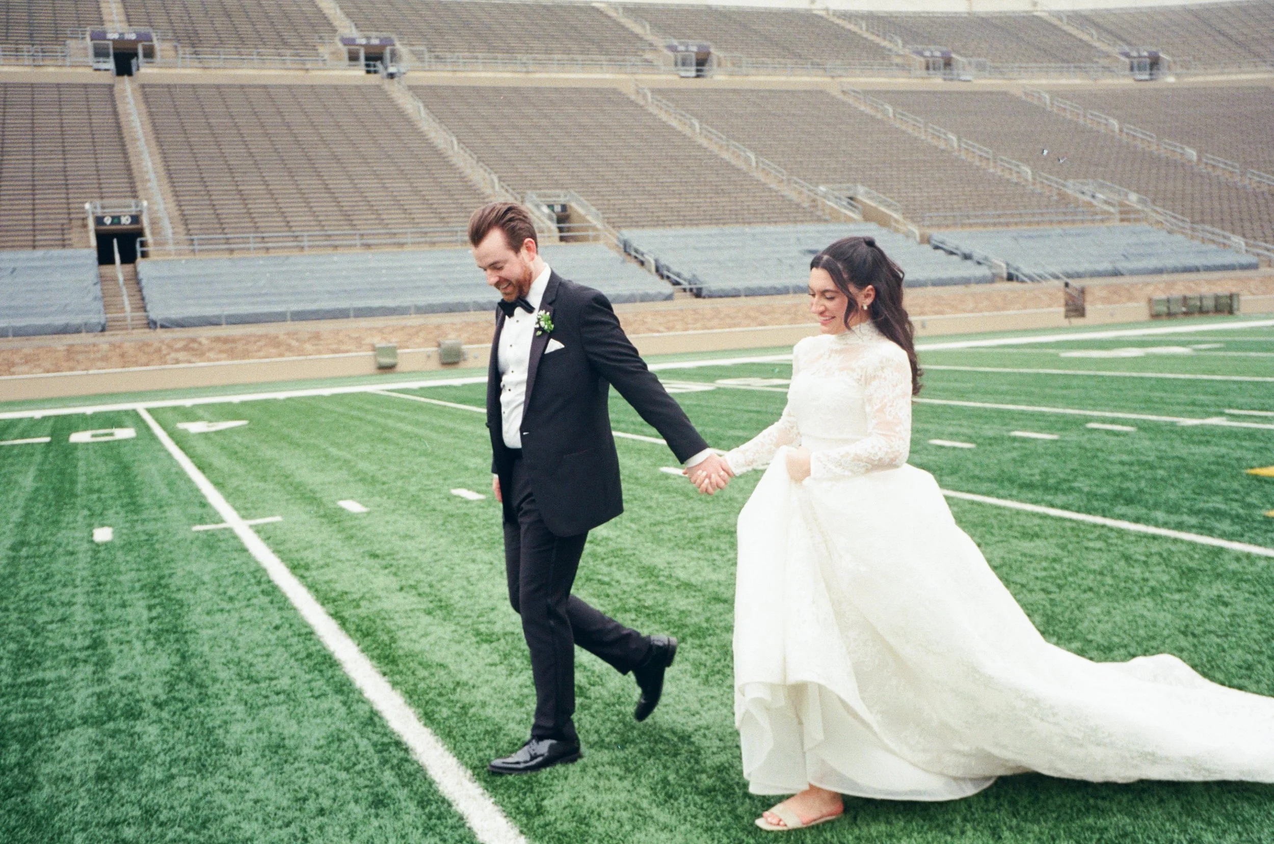 A bride and groom dressed in wedding attire holding hands on a football field inside a stadium.