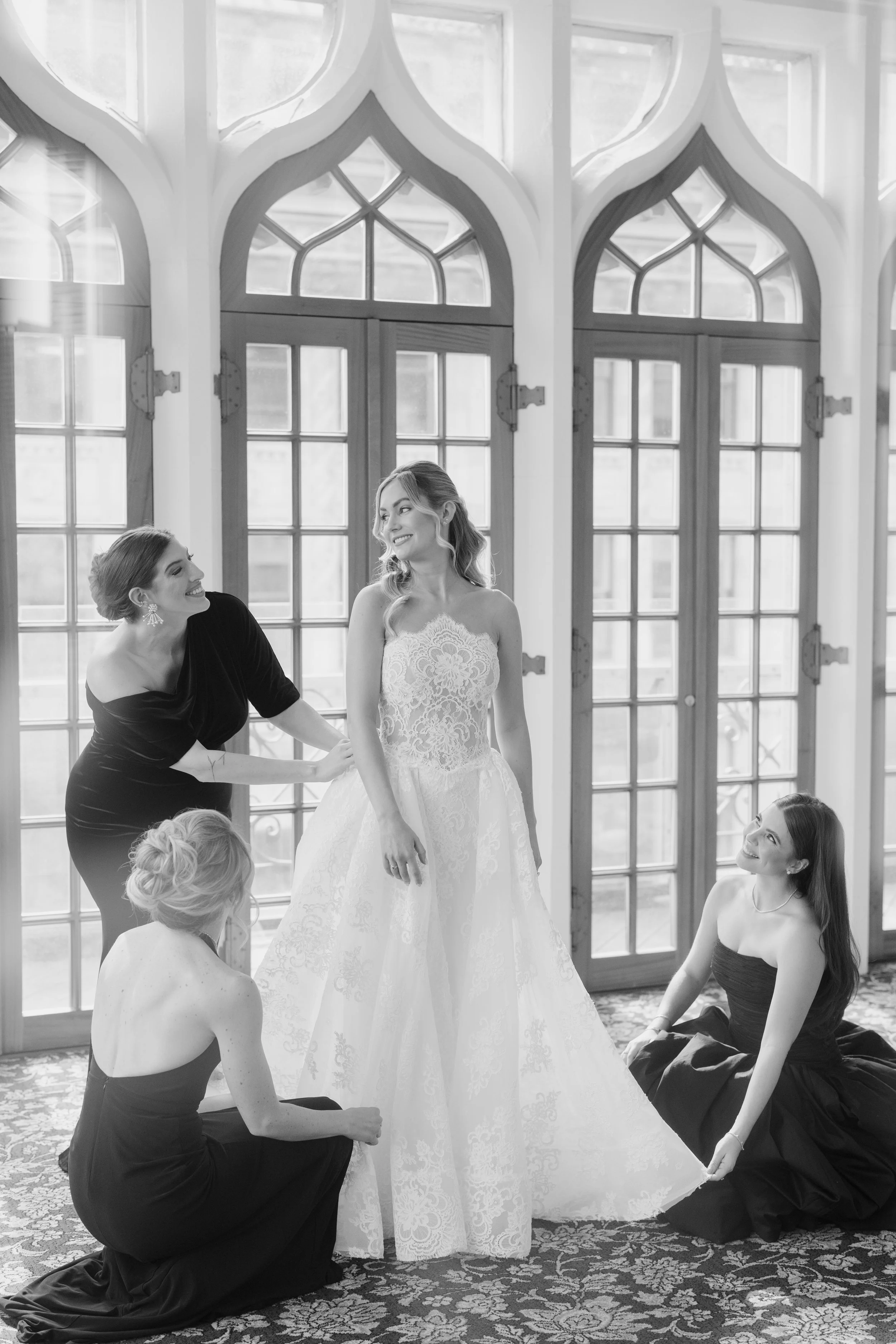 A bride in a wedding dress standing with her bridesmaids, who are helping her with her dress. The setting is a room with large decorative windows and a patterned carpet, and the women are smiling and looking at each other.
