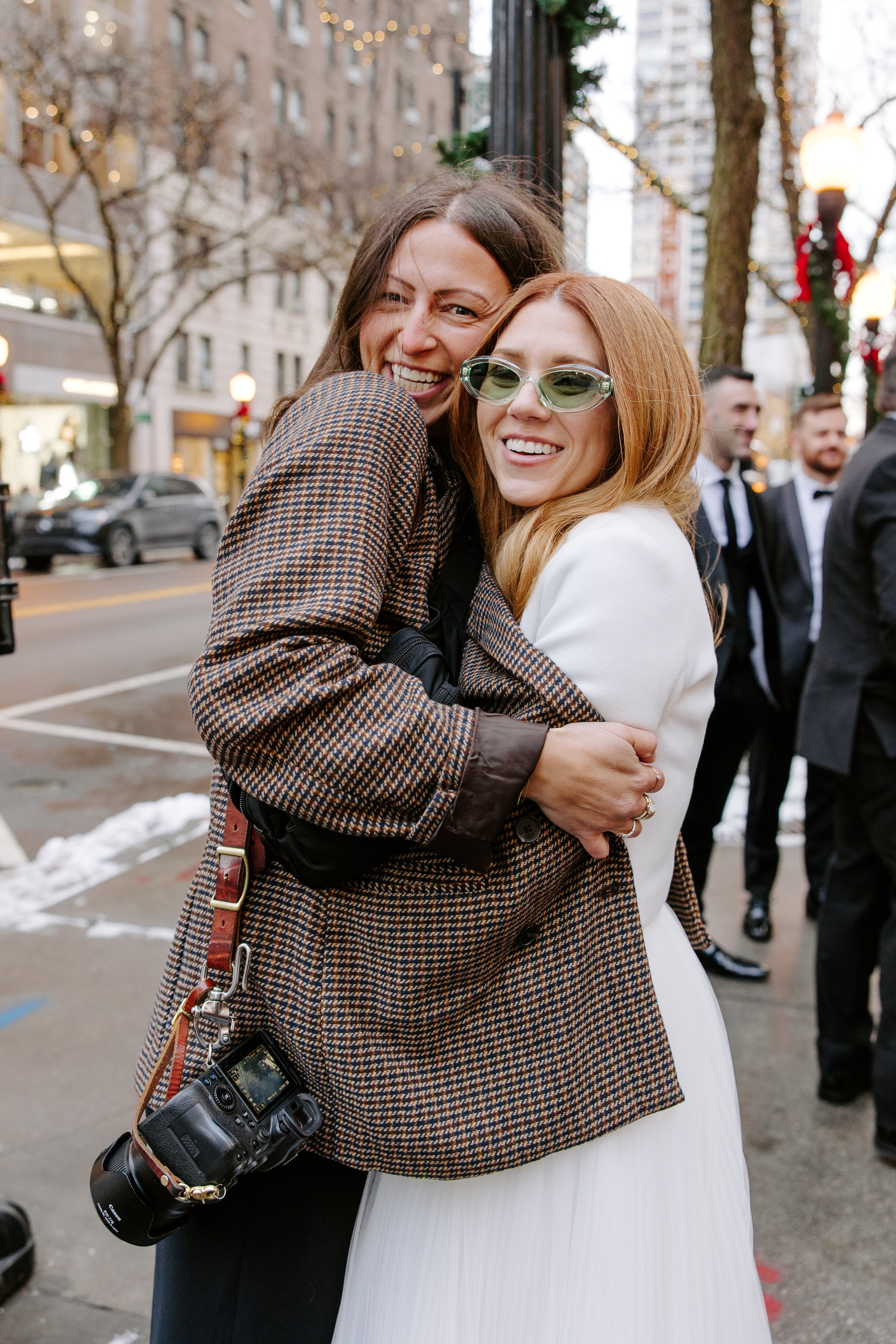 Two women embracing and smiling on a city street during winter with Christmas decorations and other people in tuxedos in the background.