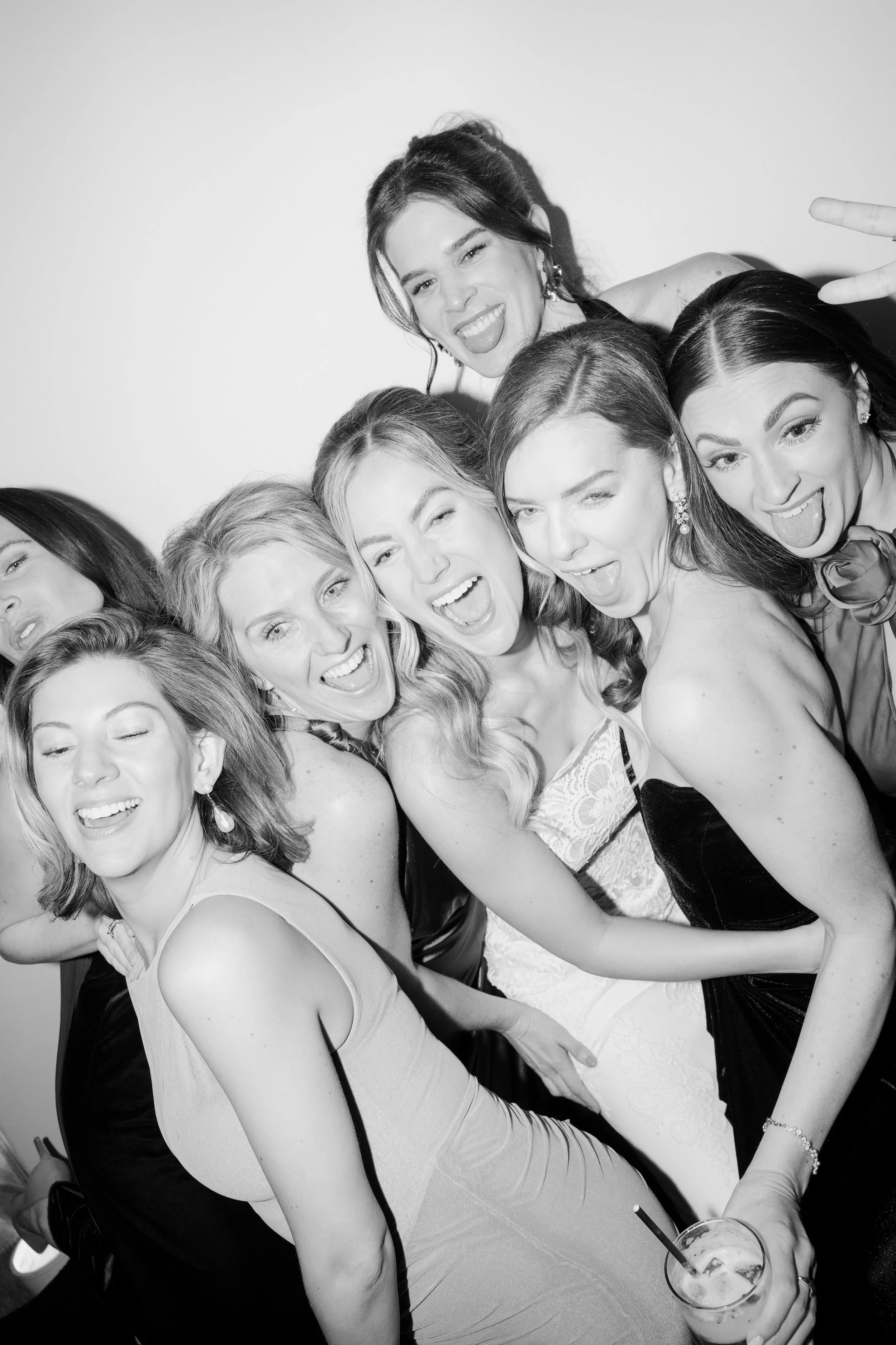 Group of women smiling and making silly faces at a party or celebration, wearing elegant dresses, with one woman holding a drink with a straw.