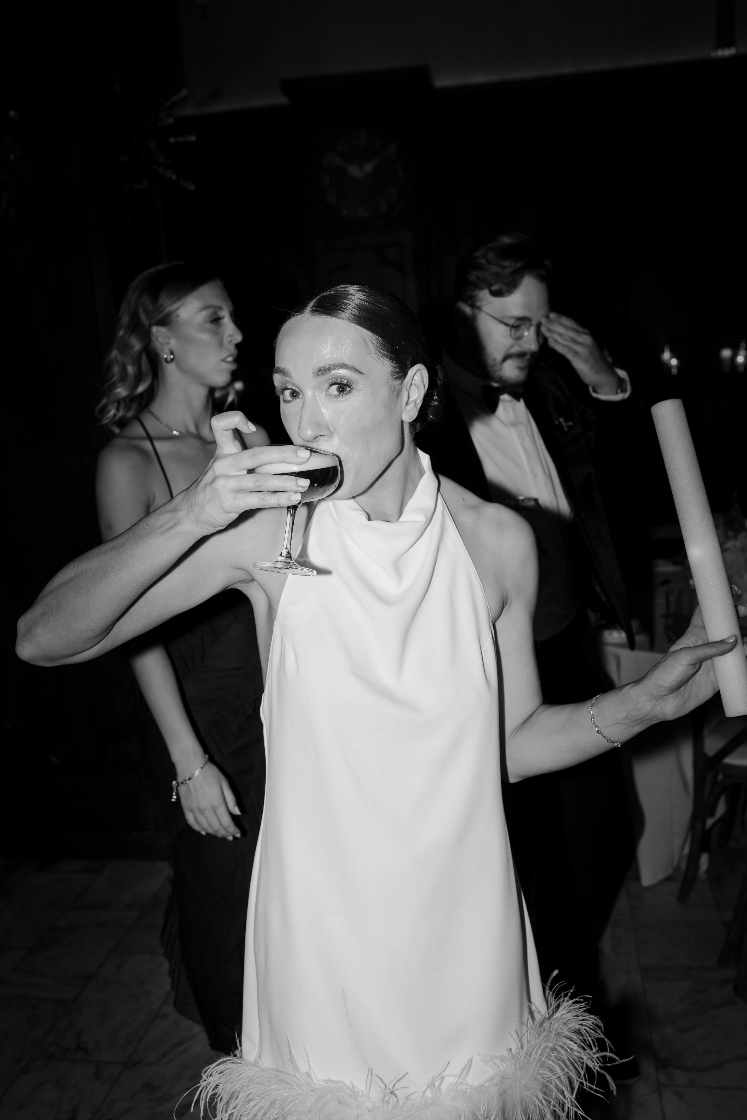 Black and white photo of a woman in a fancy dress drinking from a cocktail glass, with two other people in the background at a formal event.